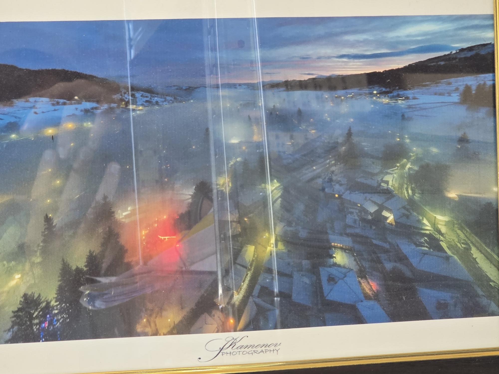 The image is a framed photograph credited to "Kamenov Photography." It depicts a cityscape or townscape, likely taken at dusk or night, with a dominant color scheme of blues, whites, and some warmer tones. The location appears to be situated in a valley or mountainous region, with hills visible on both sides of the settlement. Snow covers the ground, rooftops, and surrounding terrain, indicating a winter season. Streetlights and lights emanating from buildings punctuate the scene, adding brightness to the snowy landscape. Evergreen trees are visible, some adorned with colorful lights, suggesting a festive occasion. The scene is observed from an elevated vantage point, providing a comprehensive view of the built environment. There are no discernible people in the visible areas of the scene. The location of the city is unknown.