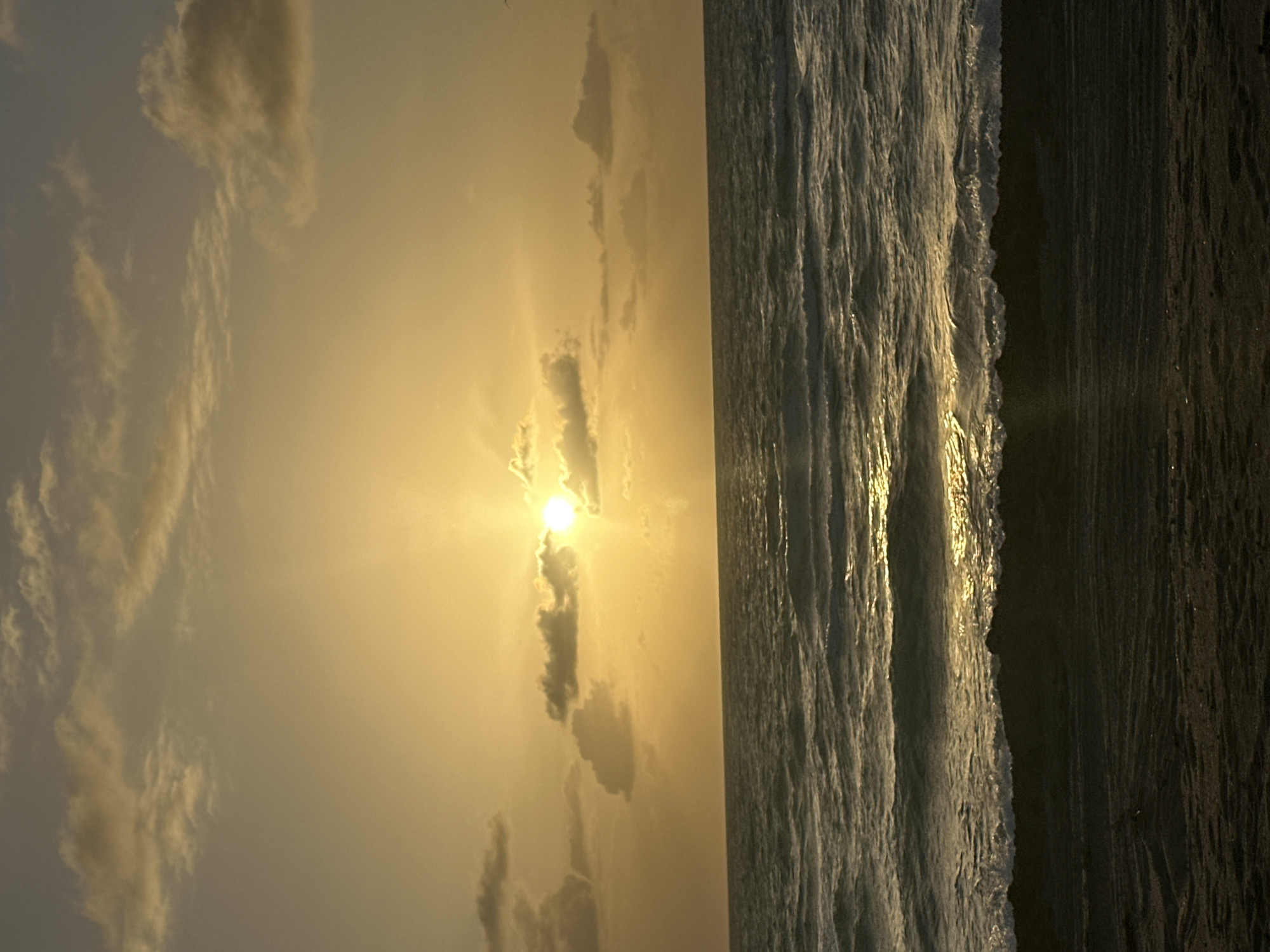 The image captures a sunset scene on Hallandale Beach, Florida, United States. The sun is positioned in the center of the frame, partially obscured by clouds, casting a golden glow across the sky and the ocean. Waves are visible crashing onto the dark sand of the beach. The overall environment is serene and indicative of a coastal sunset.