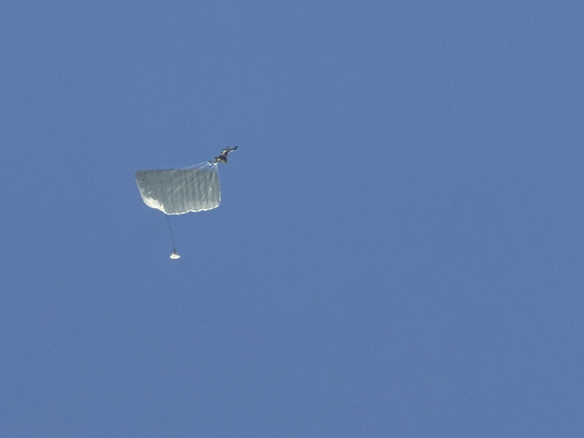 A skydiver is captured mid-air, falling against a clear blue sky. The skydiver is wearing dark clothing and is attached to a partially deployed, light-colored parachute. The parachute appears to be a rectangular canopy, with visible lines connecting it to the skydiver. The skydiver's body is positioned in a way that suggests control and stability during the descent. The image does not provide any specific location details within Korobcheevo, Russia.