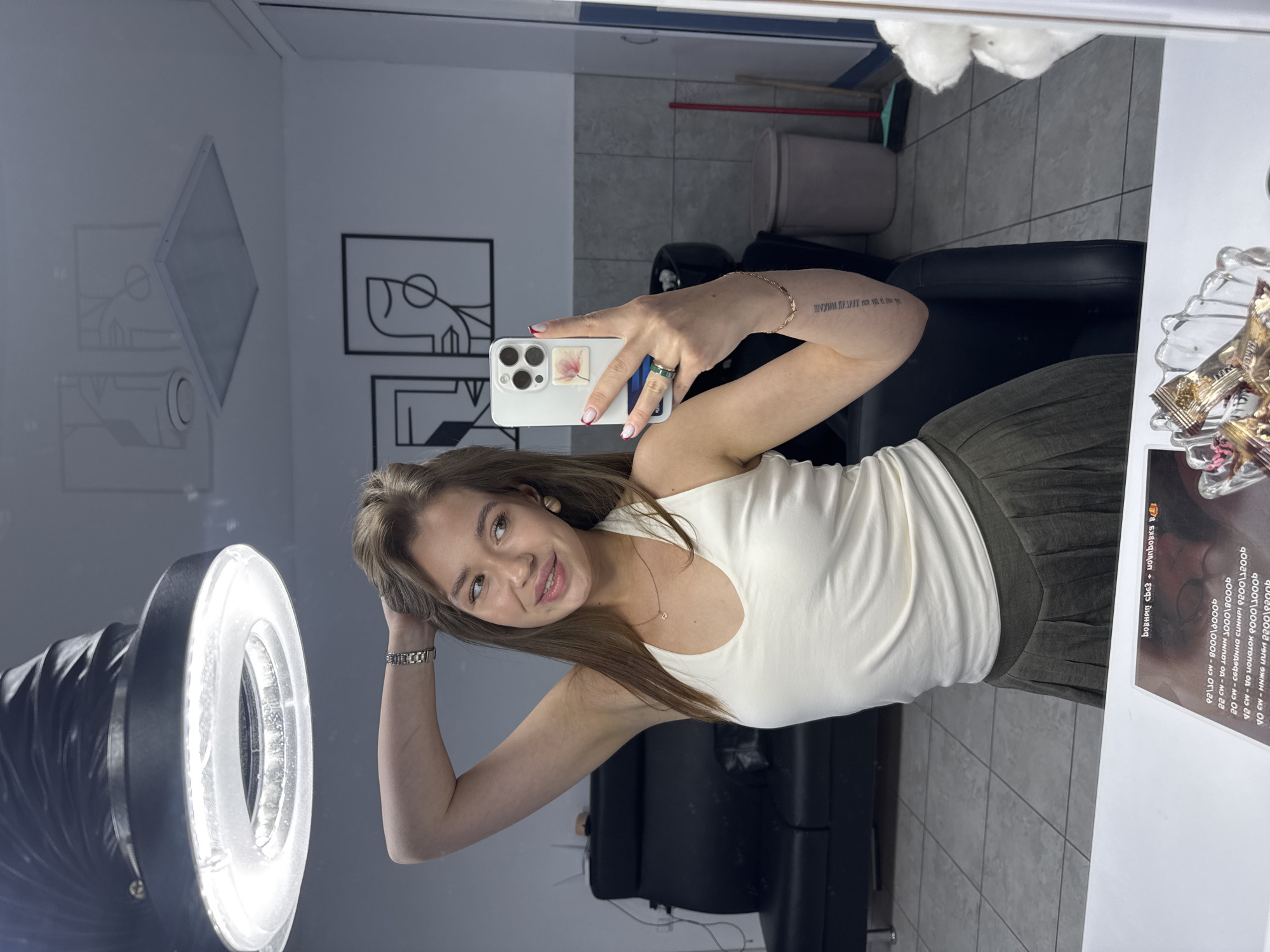 A young woman with long brown hair takes a mirror selfie. She is wearing a white halter top and a dark green pleated skirt. Her right arm is raised, with her hand resting on her head. She is looking to her right, with a slight smile. In the background, there is a black leather chair, a sink, and two framed pictures on the wall. A ring light is visible above her. The setting appears to be a salon or a similar professional space, possibly located in Chaikovsky, Russia, as indicated by the product packaging visible on the table in the foreground, which displays Cyrillic text and pricing in Russian Rubles.
