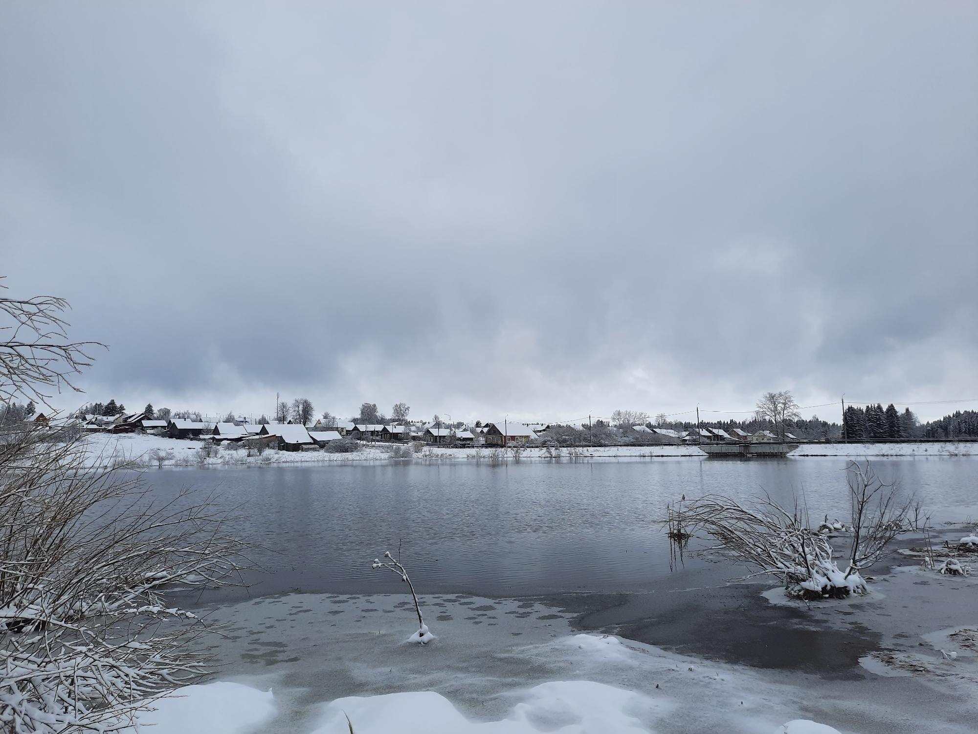 The image depicts a winter scene near Krasnogorskoye, Russia. A body of water, likely a river or lake, is partially frozen with visible ice patches and snow-covered banks. On the opposite shore, a row of houses or buildings can be seen, their roofs covered in snow. In the foreground, snow-laden bushes and frozen vegetation frame the view of the water. The sky is overcast and grey, indicating a cold, possibly snowy day. A bridge-like structure is visible in the distance, connecting the two sides of the water body. The scene is tranquil and desolate, conveying a sense of quiet isolation during the winter season.