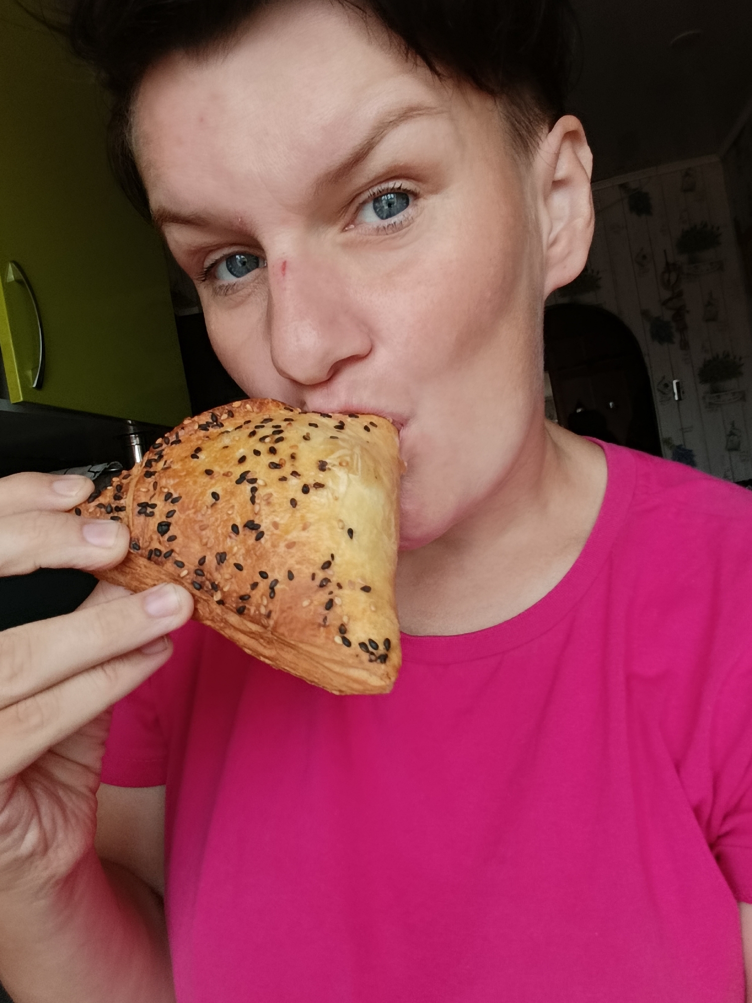 The image shows a close-up of a woman in a bright pink t-shirt eating a pastry. The woman has short dark hair and blue eyes. She is holding the pastry to her mouth with her right hand. The pastry is triangular in shape and appears to be topped with black sesame seeds. The background is blurred, but it seems to be an indoor setting. There is a hint of a green surface on the left and patterned wallpaper on the right. The image is a selfie, taken from a slightly low angle. There is no visible information that would indicate the location in the city of Фоки, Russia.