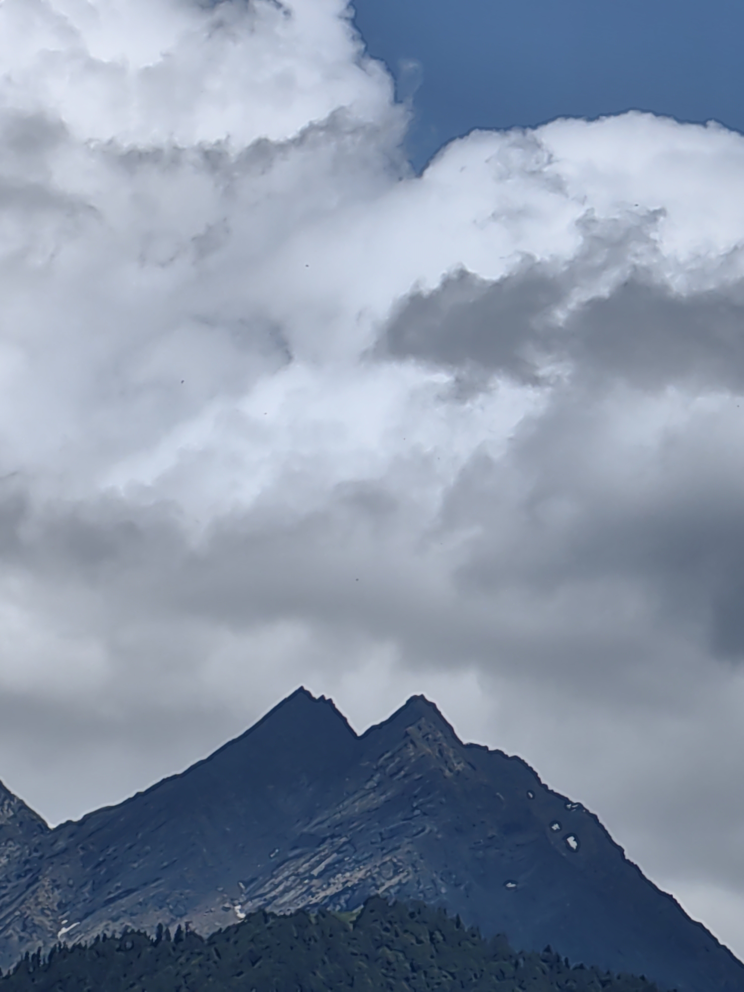 The image shows a close-up view of two jagged mountain peaks in Vashisht, India, under a cloudy sky. The mountains are dark and appear to be made of rock. There are scattered patches of snow on the slopes of the mountains. At the base of the mountains, a dense forest of dark green trees is visible. The sky is filled with a mix of white and dark gray clouds, with a small patch of blue sky visible in the upper right corner. The overall impression is one of a dramatic and majestic natural landscape.