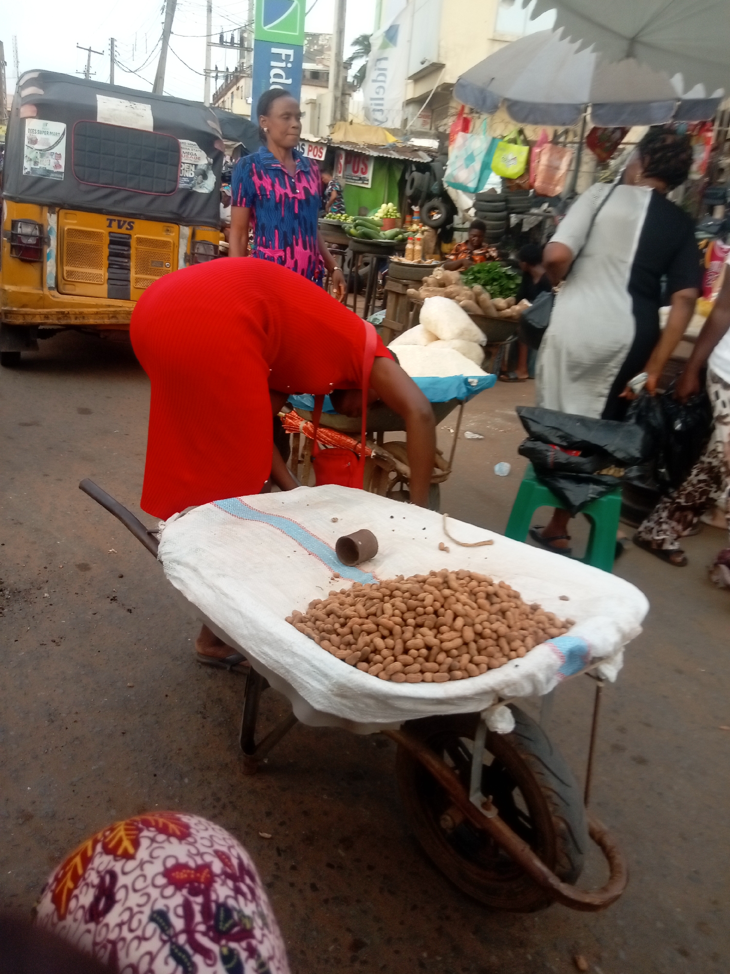 The image, taken in Nkpor, Nigeria, shows a street scene in a bustling market area. A woman in a red dress bends over a wheelbarrow full of peanuts. The wheelbarrow is covered with a white sack. A small, cylindrical metal object rests atop the peanuts. The ground is asphalt, and the foreground includes a partial view of someone's legs wearing patterned pants.
Behind the woman, various other people and objects indicate a busy market environment. A woman in a blue and pink patterned shirt stands near a yellow "TVS" tricycle. Further in the background, there are stalls selling goods, including yams, vegetables, and other items. An umbrella shades one of the stalls. People are milling about, some carrying bags. A green plastic stool is also visible in the middle ground. A sign for "Fido POS" is visible on a building in the background.