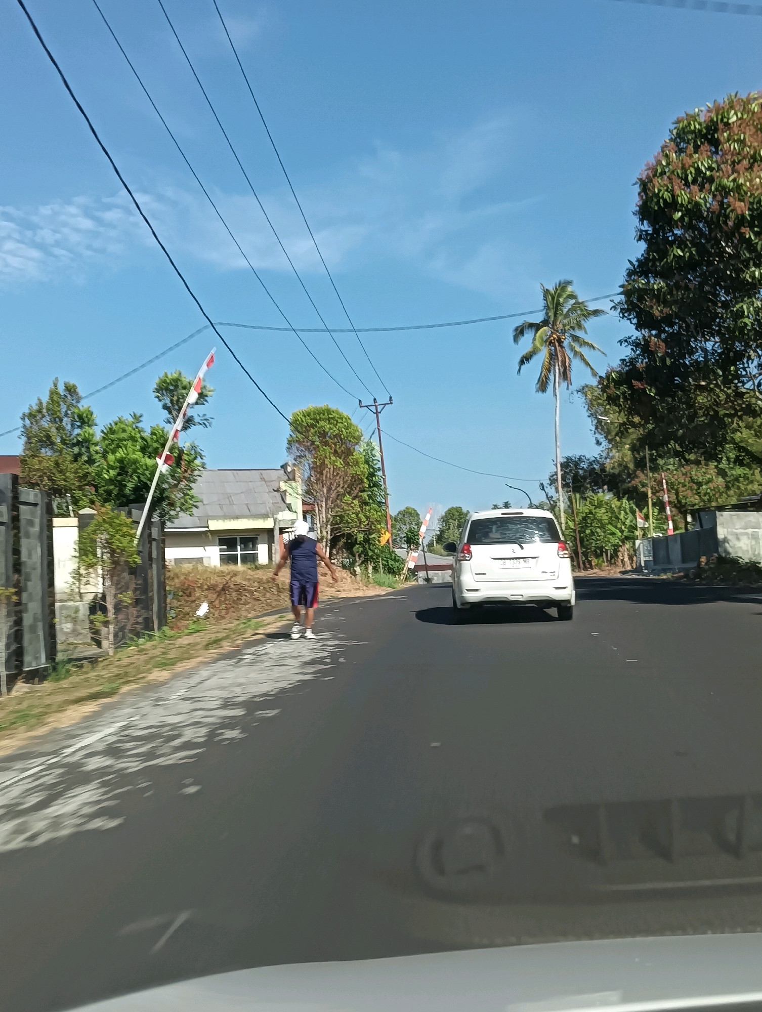 In Tomohon, Indonesia, a white car travels down a paved road under a bright blue sky. The car is the dominant object in the foreground, with a person walking on the side of the road in the middle ground. The person is wearing a dark sleeveless shirt and blue shorts with red trim, and is facing away from the camera. Next to the person is a red and white flag on a pole, and a building with a grey roof. To the right of the road, there is a palm tree and other foliage. The road is lined with a curb and a grassy shoulder. The overall scene is of a typical Indonesian street scene on a sunny day.