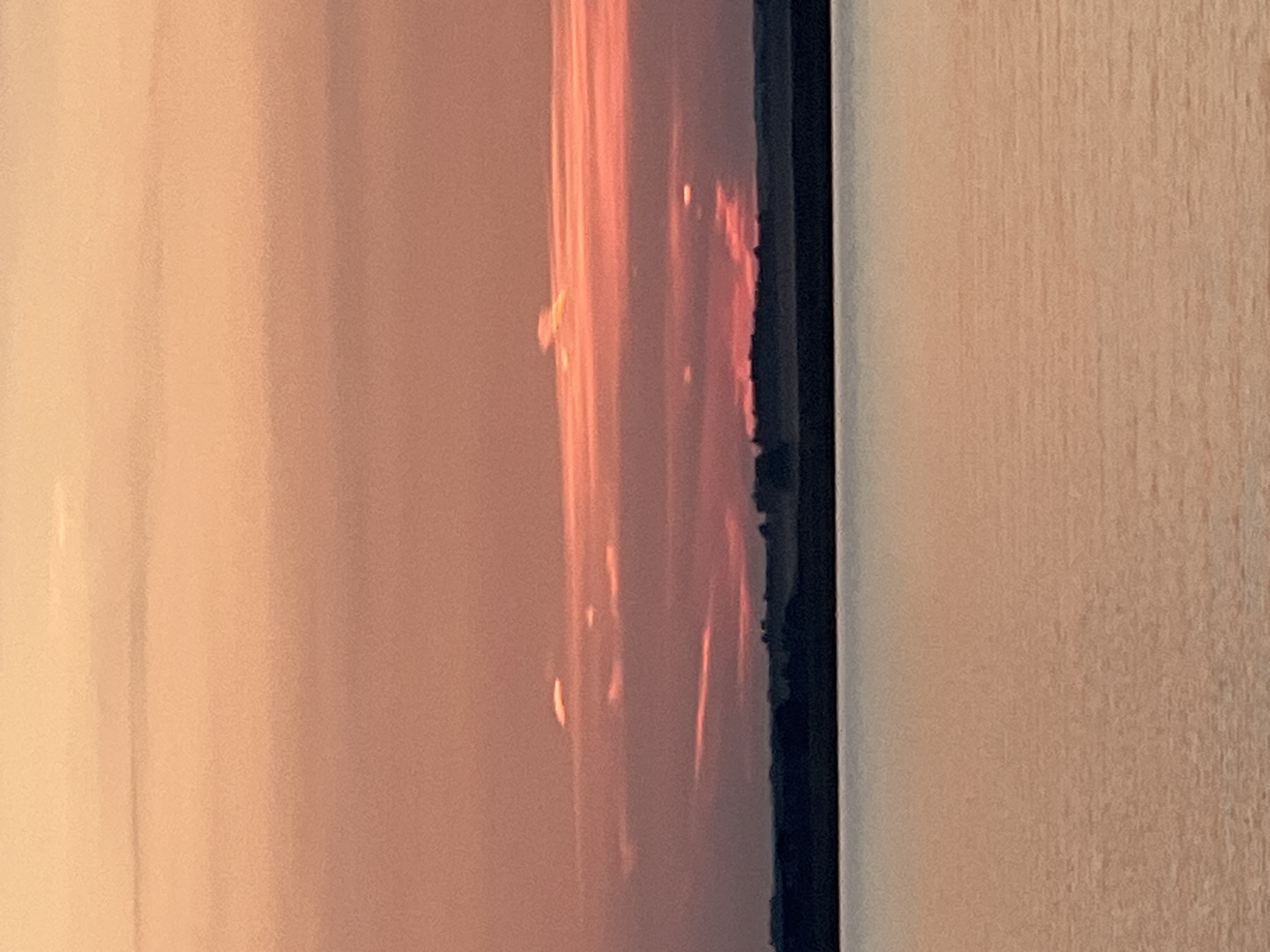 A sunset over a body of water is captured in a horizontal, close-up shot. The sky is a gradient of soft oranges and pinks, with thin bands of clouds stretching across the frame. The clouds are tinged with a brighter coral color where they catch the last rays of the sun, with some appearing as small, luminous streaks. Below the sky, a dark, silhouetted landmass is visible, suggesting a distant shore or a line of trees. The water in the foreground reflects the colors of the sky, creating a muted, hazy surface with subtle horizontal ripples. The overall impression is one of serene tranquility. This scene depicts the natural environment of Novopichugovo, Russia, during twilight.