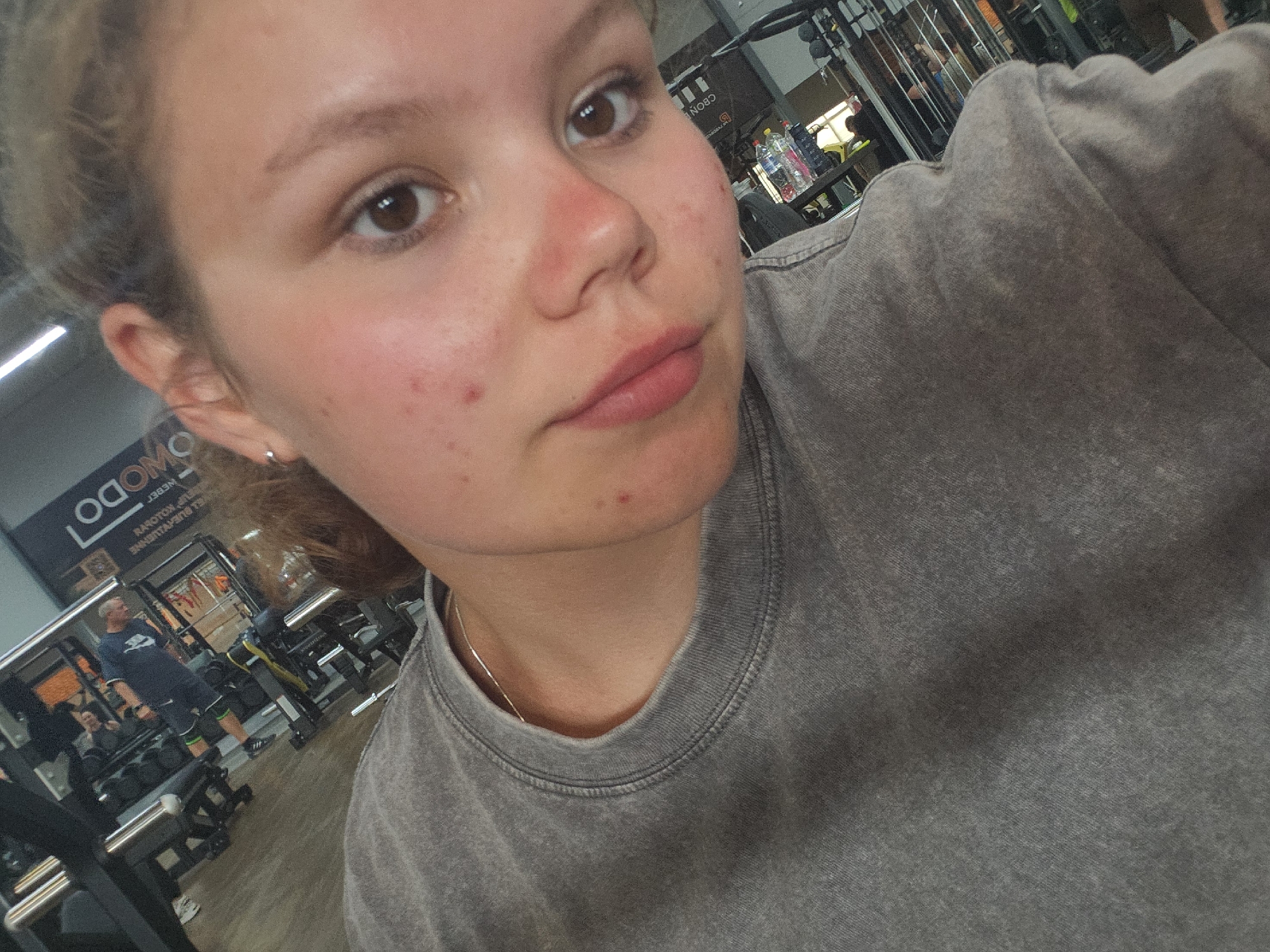 The image shows a close-up, angled selfie of a young person in a gym. The person has light skin with some redness and acne on their face. They are wearing a gray sweatshirt. In the background, gym equipment, including weight machines and racks of weights, is visible. There are other people in the gym, but they are out of focus and not clearly identifiable. The overall environment appears to be a modern fitness facility. The presence of a sign with Cyrillic lettering, specifically "MODO," suggests a Russian-speaking country. Chaikovsky is a city in Russia known for its sports facilities, making a gym setting plausible.