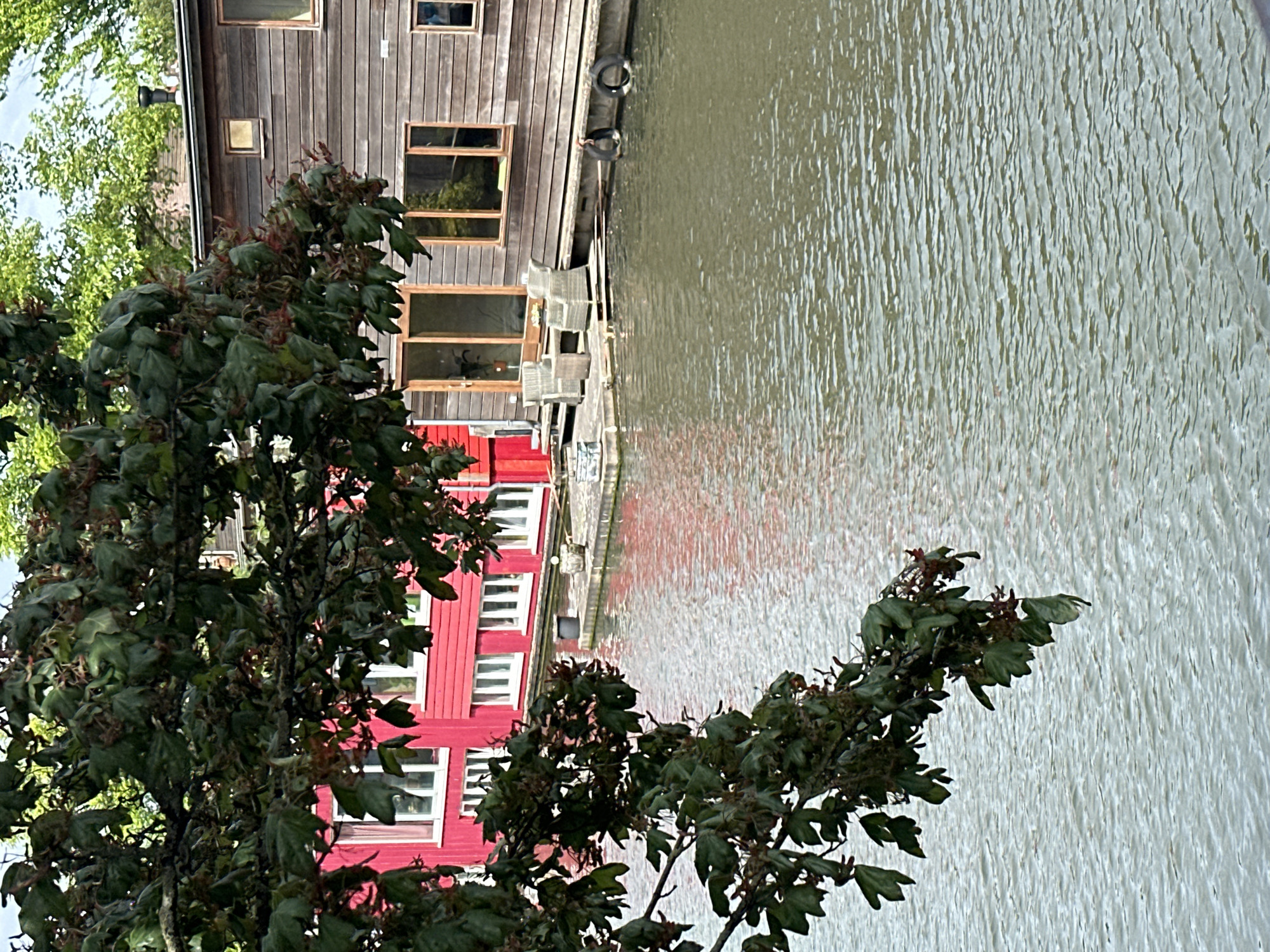 The image depicts a scene along a canal in Amsterdam, Netherlands, with a house boat moored along the water's edge. The boat's structure includes a weathered wooden upper section and a brightly painted red lower section with windows. A tree with dense foliage partially obscures the view of the house boat, adding depth to the composition. The canal water reflects the house boat and the red structure of the lower section. Patio furniture is visible on the deck of the house boat.