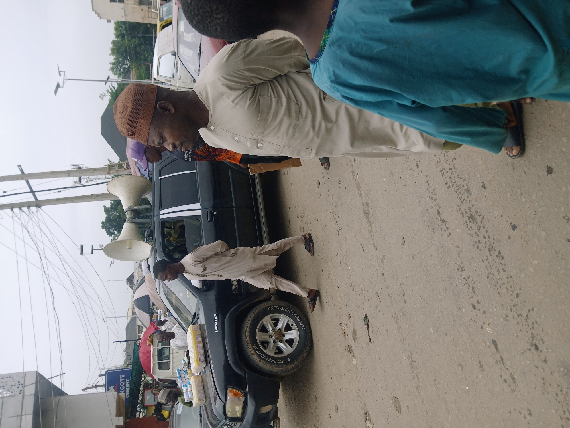 The image shows a street scene in Abuja, Nigeria. A black SUV is parked on the side of the road with loudspeakers mounted on its roof. There are people gathered around the vehicle. A man wearing a brown kufi and a light-colored traditional outfit is standing near the SUV. Another man in blue clothing is partially visible in the foreground. Various items, possibly for sale, are displayed on the front of the SUV. A sign indicating "ANGOTE CEMENT" is visible in the background. Power lines run overhead. The sky appears overcast.