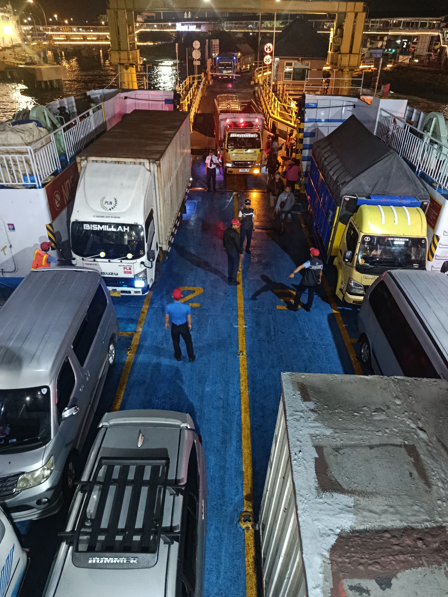 An aerial, high-angle, night-time, medium shot captures a ferry deck filled with vehicles and people. The ferry is docked at the port in Melaya, Indonesia. The deck is painted blue with yellow lines. In the foreground, a gray SUV with a roof rack is parked. To its left, a gray minivan is parked. Further back, a white truck with a container is parked, and to its right is a yellow truck with a blue cargo cover. Several individuals, some in uniform, are directing traffic or observing the scene. The background shows port infrastructure, including lights and railings, with a body of water visible. The overall impression is one of a busy, organized loading or unloading operation.