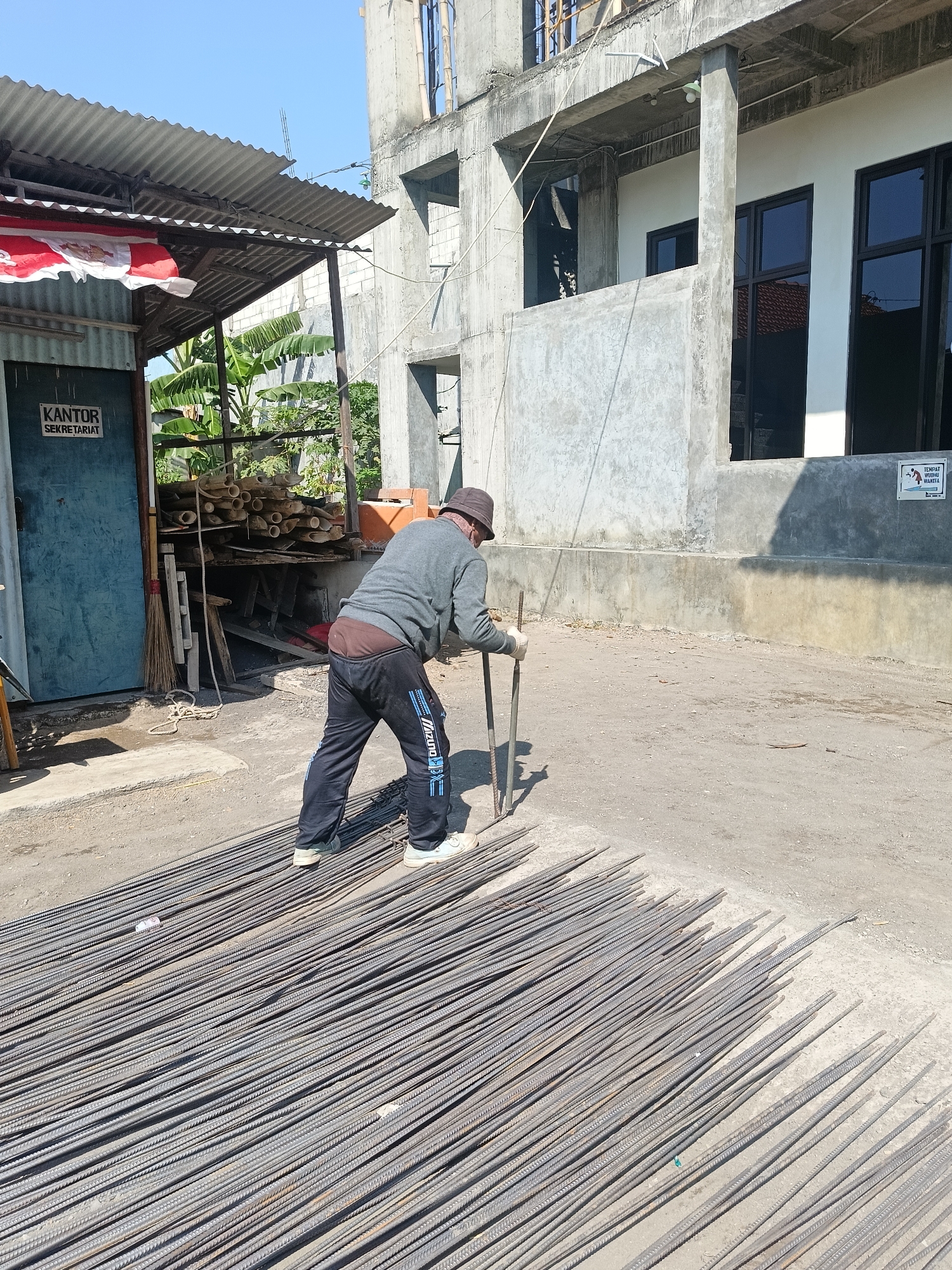 A man wearing a hat, a grey long-sleeved shirt, and dark pants with blue stripes is standing on a pile of rebar rods. He is holding two rebar rods vertically in his hands, appearing to be working with them. The scene takes place outdoors in what appears to be a construction area in Gedangan, Indonesia. To the left of the man is a blue door with a sign that reads "KANTOR SEKRETARIAT" (Office Secretariat). Behind the door, there is a sheltered area with corrugated metal roofing and stacked bamboo poles. In the background, a partially constructed concrete building with windows is visible. The ground is a mix of dirt and concrete.