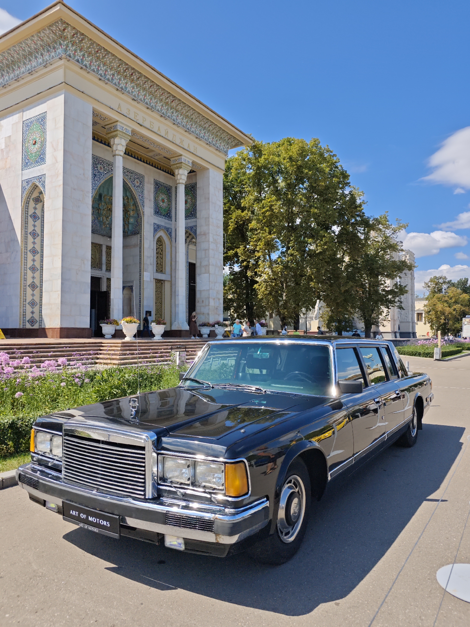 A black, four-door limousine, likely a ZIL, is parked on an asphalt surface in front of a white building with ornate blue and green tilework and a gold inscription. The building has large columns and steps leading up to an entrance. Several potted plants are arranged on the steps. Green trees and a blue sky with scattered clouds are visible in the background. Several people are present in front of the building and further in the background. A license plate on the front of the car reads "ART OF MOTORS". The inscription on the building appears to read "АЗЕРБАЙДЖАН", suggesting the building is the Azerbaijan pavilion. This location is likely at the All-Russian Exhibition Centre (VDNKh) in Moscow, Russia, which historically housed pavilions for various Soviet republics.