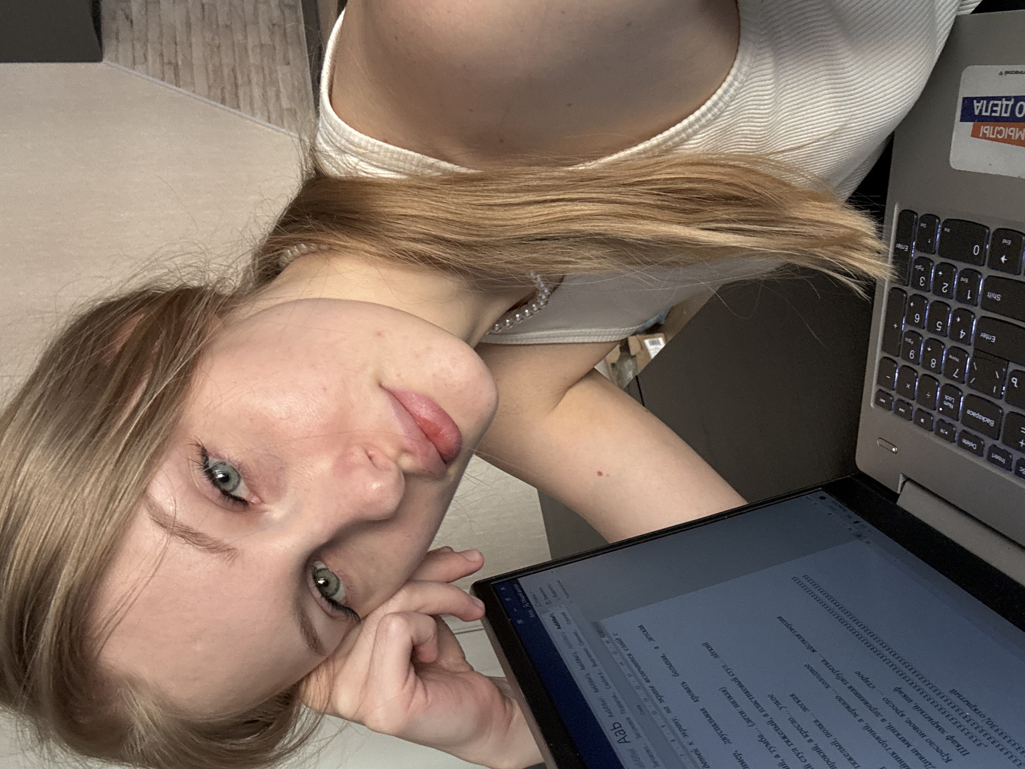 The image presents a young woman captured from an overhead perspective, looking directly at the camera with a relaxed, slightly smiling expression. She has light-colored hair, which cascades around her head, and striking blue-green eyes. She wears a white ribbed tank top, and a pearl necklace is visible around her neck. Her head is resting on her hand, indicating a moment of comfort or pause.
The scene is an indoor setting, likely a home or workspace in Perm, Russia, as suggested by the visible text cues. To her right, a silver laptop with a keyboard displaying both English and Cyrillic characters is open. A sticker on the laptop lid reads "АМБИЦИИ И ДЕЛА" (Ambitions and Affairs/Business), "МЫСЛЫ" (Thoughts), and partially "ПЕРМЬ" (Perm), reinforcing the location. In front of her, an additional screen or tablet displays a document primarily in Cyrillic text, suggesting she might be working, studying, or engaged in research.
Visible text on the screen includes formatting options like "Заголовок" (Heading) and "Подзаголовок" (Subheading), along with several lines comparing opposing concepts, such as "Кресло новое, кресло... старое" (new chair, old chair), "Шкаф закрытый, шкаф... открытый" (closed closet, open closet), "Диван мягкий, а деревянная табуретка... жёсткая твердая" (soft sofa, hard wooden stool), and "чайник горячий, а зеркало... холодное" (hot kettle, cold mirror). There's also a phrase "детей к экрану" (children to the screen) and repetitive sequences of "33333". The background shows a light-colored surface, possibly a carpet, next to wooden floorboards. The lighting appears natural, suggesting it is daytime.