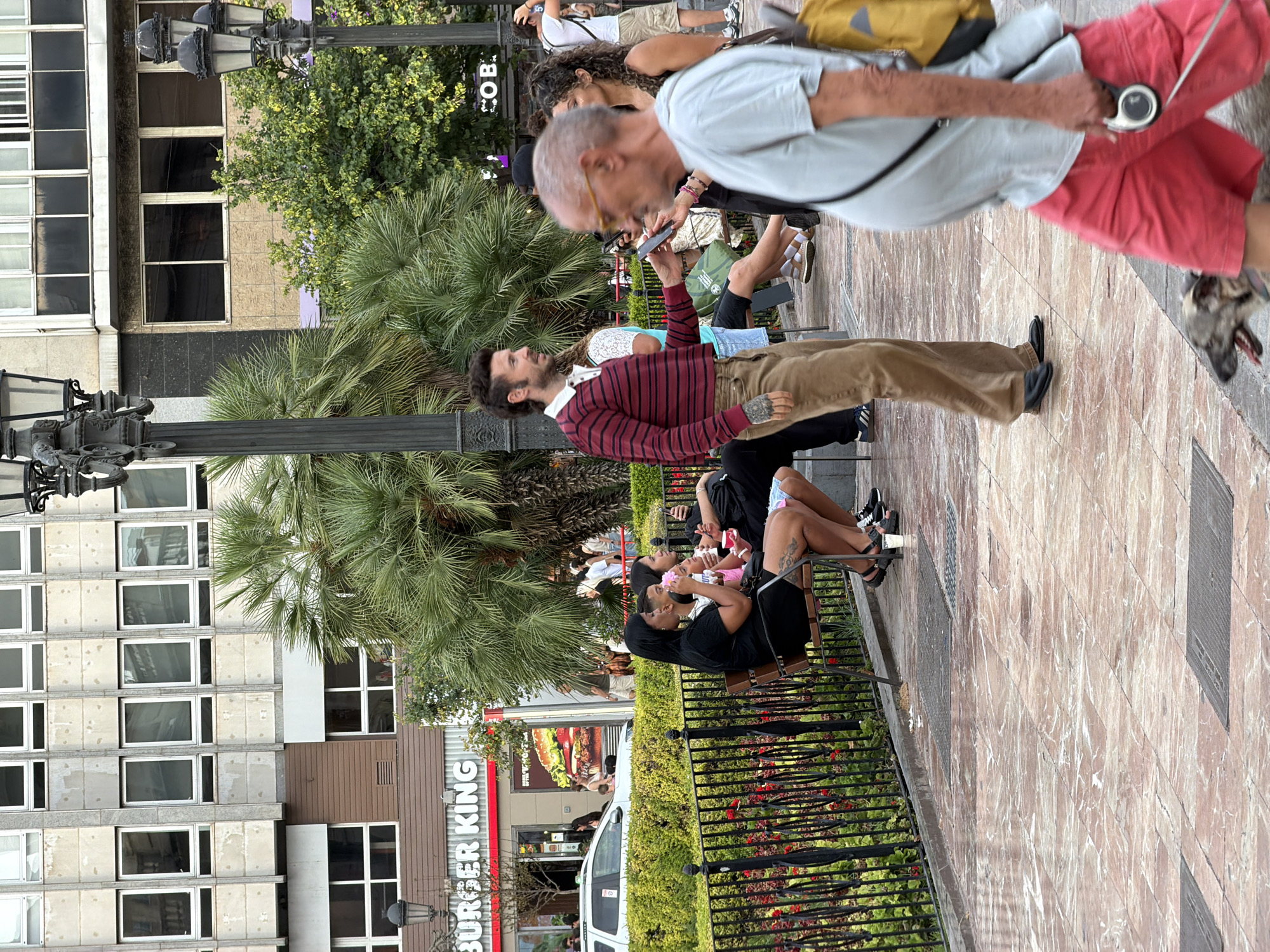 The image depicts an outdoor urban scene, possibly a plaza or pedestrian area. In the foreground, an older man with glasses, wearing a light blue t-shirt and red shorts, walks a dog on a leash. He has a yellow backpack slung over his shoulder. To his left, a man with a mullet hairstyle and a striped long-sleeve shirt is interacting with someone out of frame, holding a phone. He is wearing brown pants and black shoes. Behind him, a group of people are seated on a bench, some holding food and drinks, with a black metal fence and greenery behind them. In the background, a Burger King restaurant is visible, as well as a multi-story building with numerous windows. Several palm trees and other foliage are interspersed throughout the scene. The ground is paved with marble-like tiles.