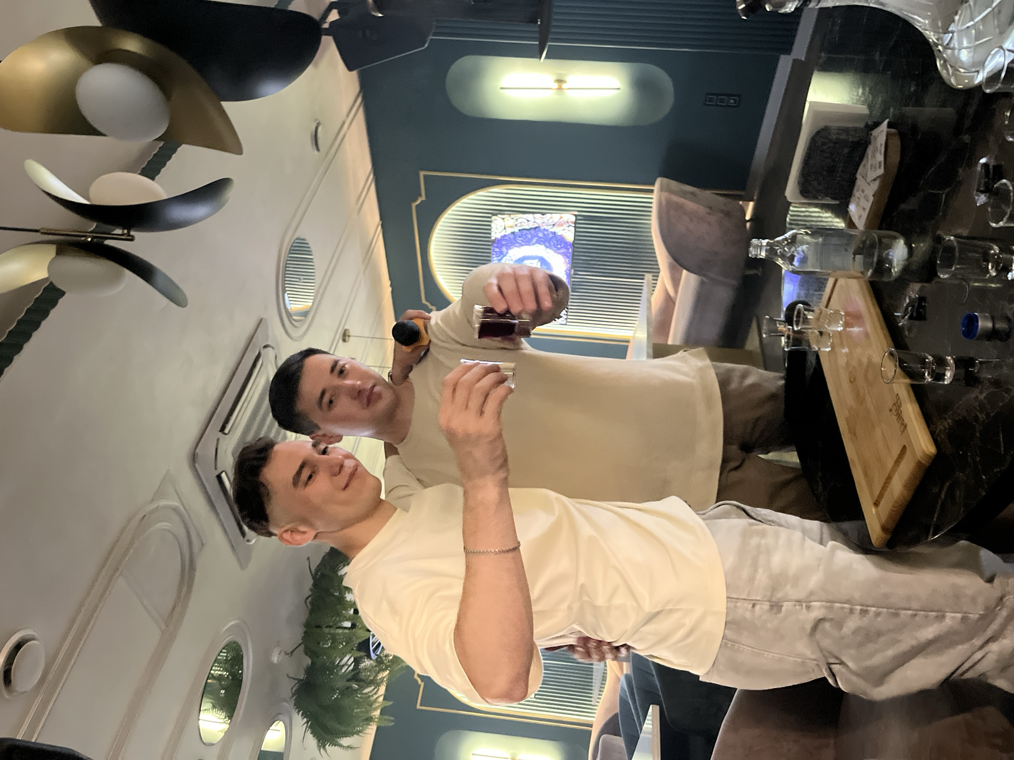 The image shows two young men standing in a dimly lit bar or restaurant with a modern interior. The man on the left, wearing a light-colored t-shirt and light-colored pants, is holding up a shot glass with a dark liquid and smiling. The man on the right, wearing a similar light-colored t-shirt, is also holding up a shot glass and looking towards the camera. They are standing in front of a dark, reflective table on which several other shot glasses, a wooden board, and a bottle of clear liquid are visible. In the background, there is a dark teal wall with decorative gold accents and an illuminated arched feature. The ceiling has modern light fixtures and ventilation. The overall atmosphere suggests a casual gathering or celebration. The image does not provide any specific visual cues to pinpoint the location within Naberezhnye Chelny, Russia.