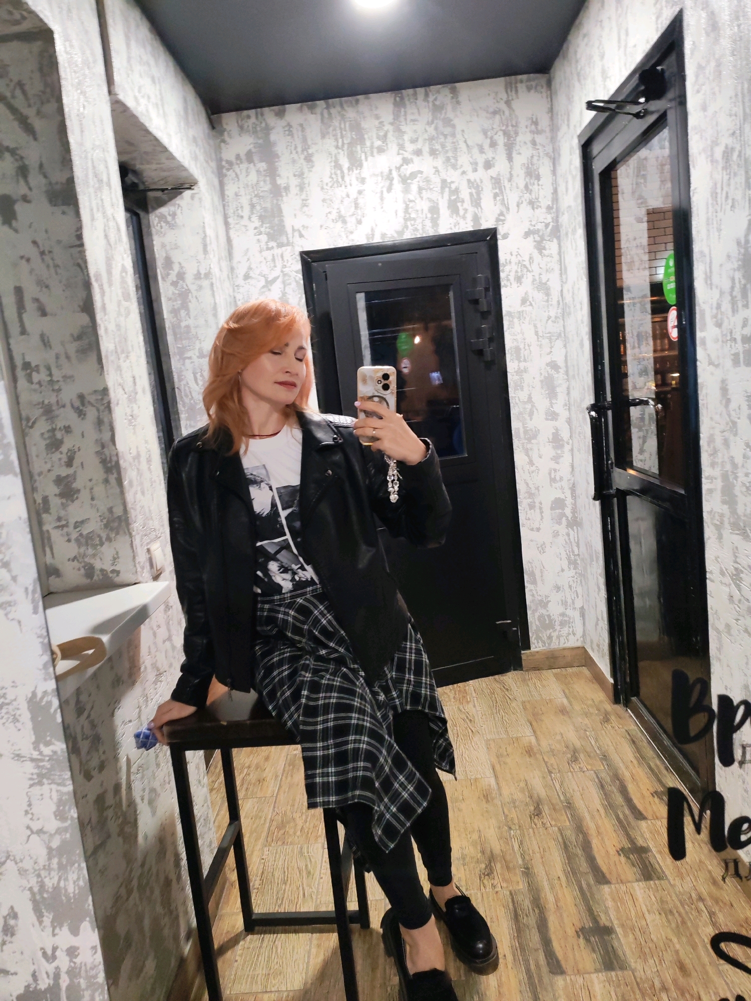 A woman with reddish-blonde hair takes a mirror selfie in a hallway with textured grey and white walls. She is wearing a black leather jacket over a white t-shirt with a black and white graphic print and a black and white plaid skirt. She is also wearing black leggings and black loafers. She is sitting on a tall, black stool. A door is visible behind her, with a glass pane showing a reflection of what appears to be a shop interior. The floor is made of light brown wood-look laminate. The woman is holding a smartphone in her right hand. The scene is likely indoors, possibly in a commercial establishment or a residential hallway in Chaikovsky, Russia, given the presence of a door and the overall interior design.