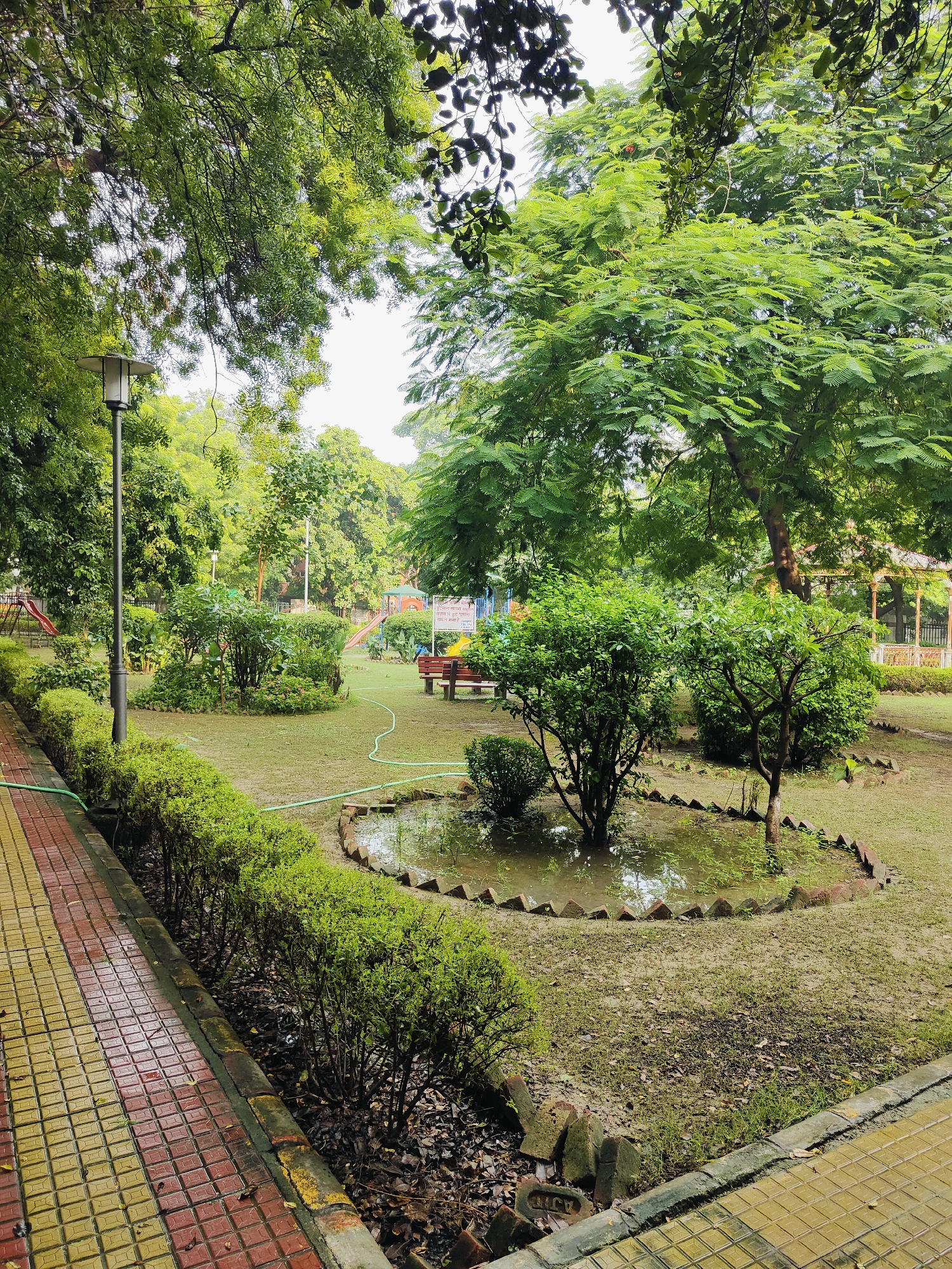 The image depicts a park in Gurgaon, India, likely during or after rain. A paved walkway with yellow and red tiles runs along the left side of the frame, bordered by manicured green bushes. Beyond the bushes, a grassy area contains a small, flooded garden bed with a circular brick border and a young tree. In the background, a park bench is visible, along with playground equipment including slides. The park is surrounded by lush green trees, indicating a well-maintained urban green space. A lamppost stands near the walkway. The overall atmosphere is peaceful and verdant.