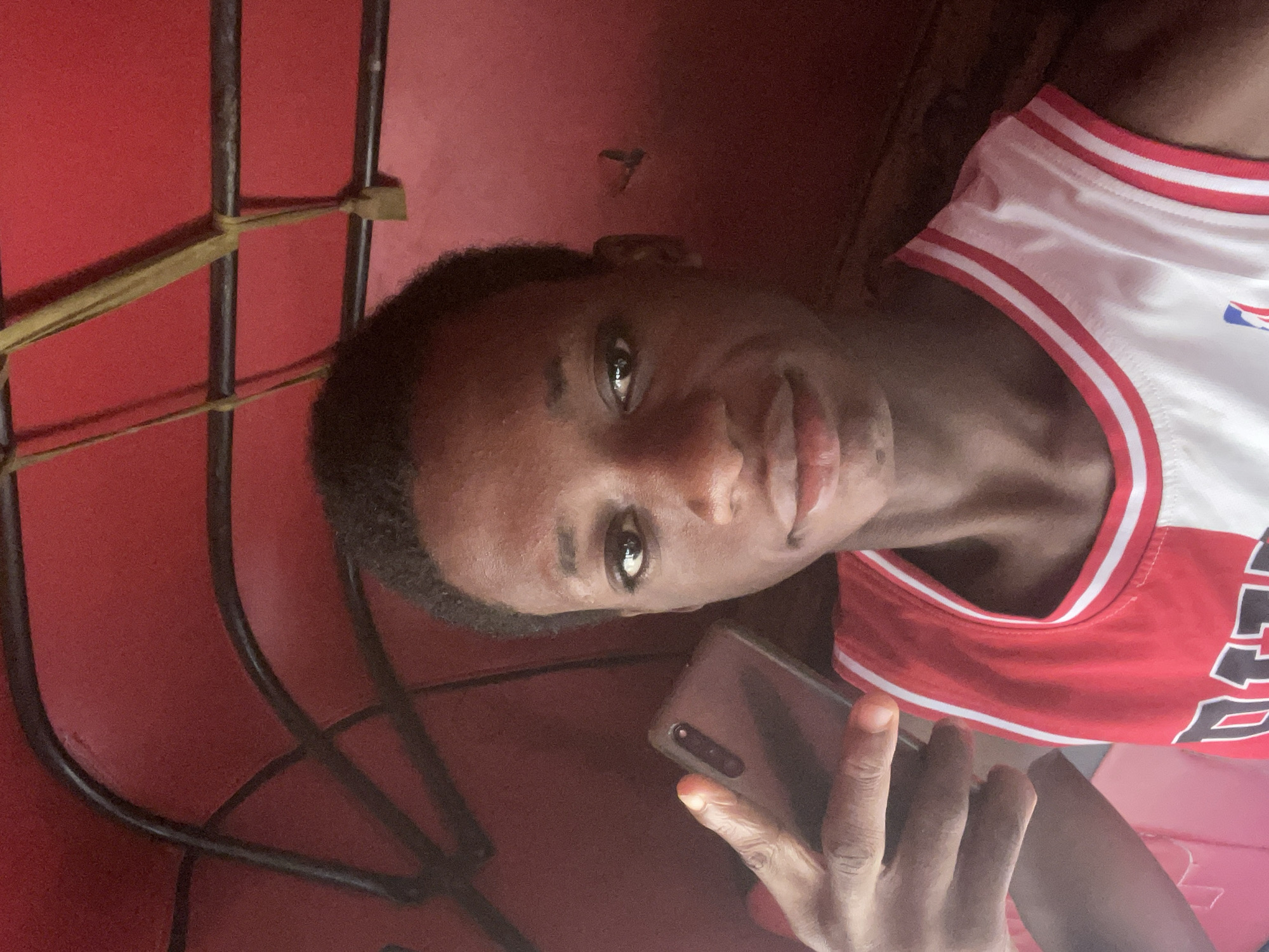 A young man is taking a selfie inside an auto rickshaw (keke) in Onitsha, Nigeria. The keke has a red interior and black metal safety bars secured with yellow ties. The man is wearing a red and white basketball jersey and holding a smartphone.
