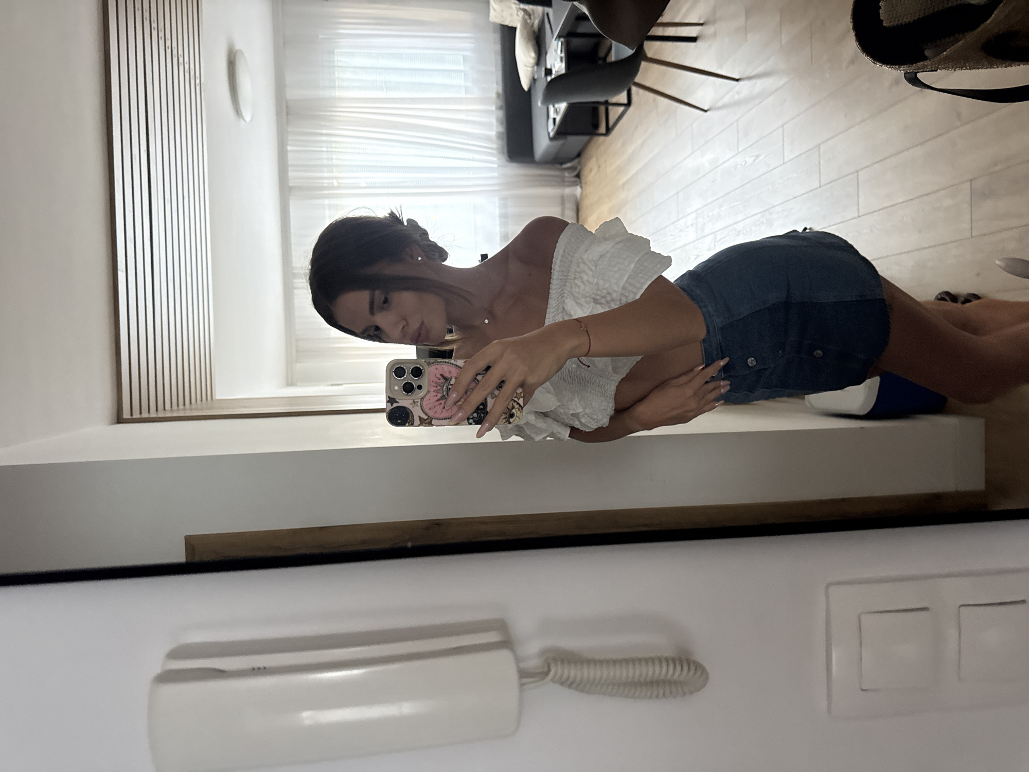 A young woman with dark hair styled in a bun is taking a mirror selfie in what appears to be a residential apartment. She is wearing a white off-the-shoulder top with ruffled details and a dark denim skirt. Her left hand rests on her hip, and she is holding a smartphone with a decorative case in her right hand. The background of the reflection shows a living area with a sofa, coffee table, and chairs, as well as sheer curtains covering a window. The room has light-colored wooden flooring. To the left of the woman, in the foreground of the actual scene, is a white intercom system mounted on a white wall, and a portion of a doorway with a wooden frame. The overall impression is that of a casual, indoor setting. The image does not contain any discernible elements that would indicate its specific location within the city of Plovdiv, Bulgaria.