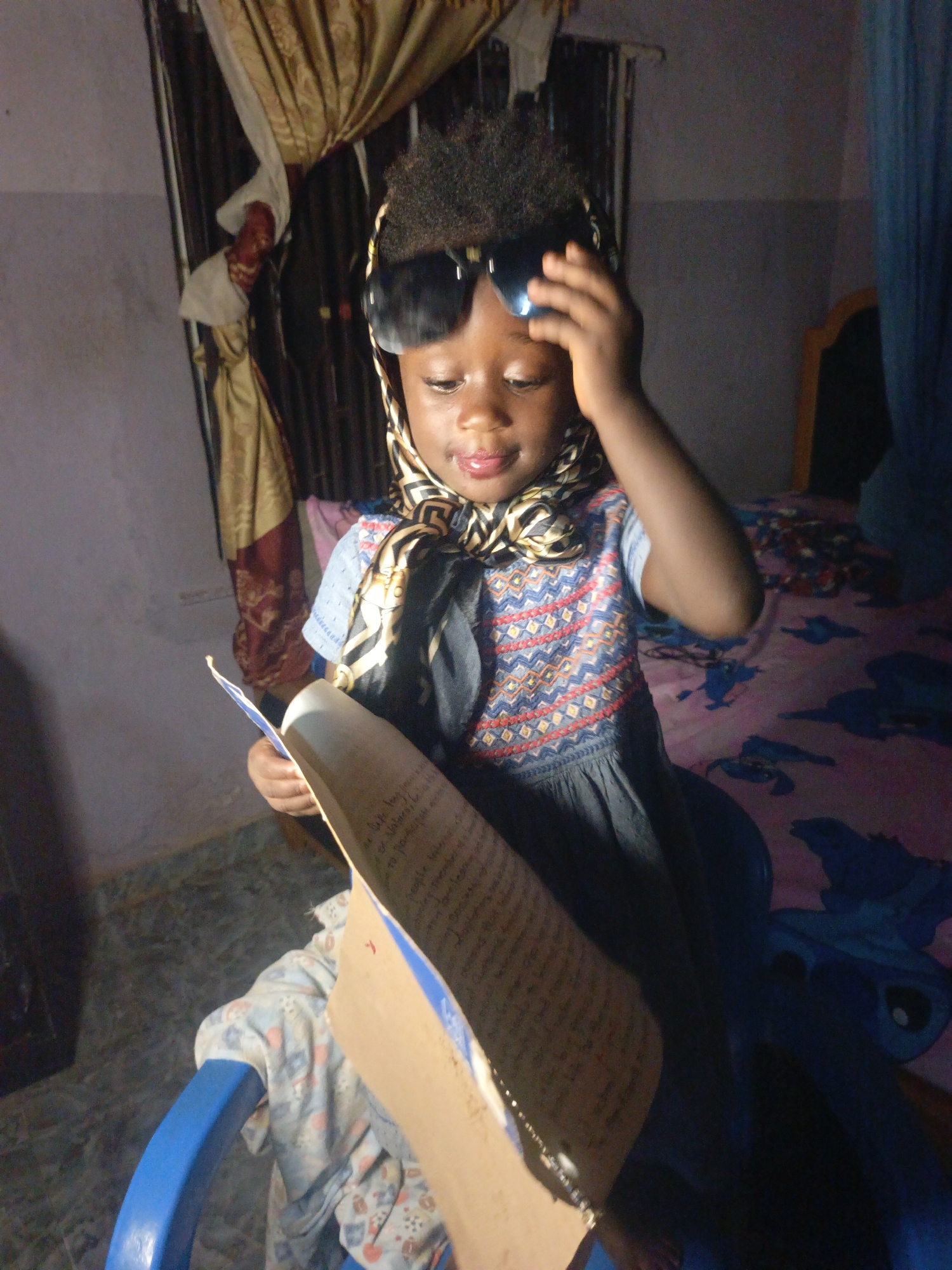 A young child is seated indoors, likely in a home, potentially in Akwanga, Nigeria given the prompt. The child is wearing sunglasses on their head and a patterned scarf around their neck. They are holding an open notebook or folder with handwritten notes. A blue child's chair is beneath them, covered with a patterned cloth. Behind the child, there are curtains and a bed with a blue patterned sheet. The walls are painted a light color. The child is touching their sunglasses with their right hand.