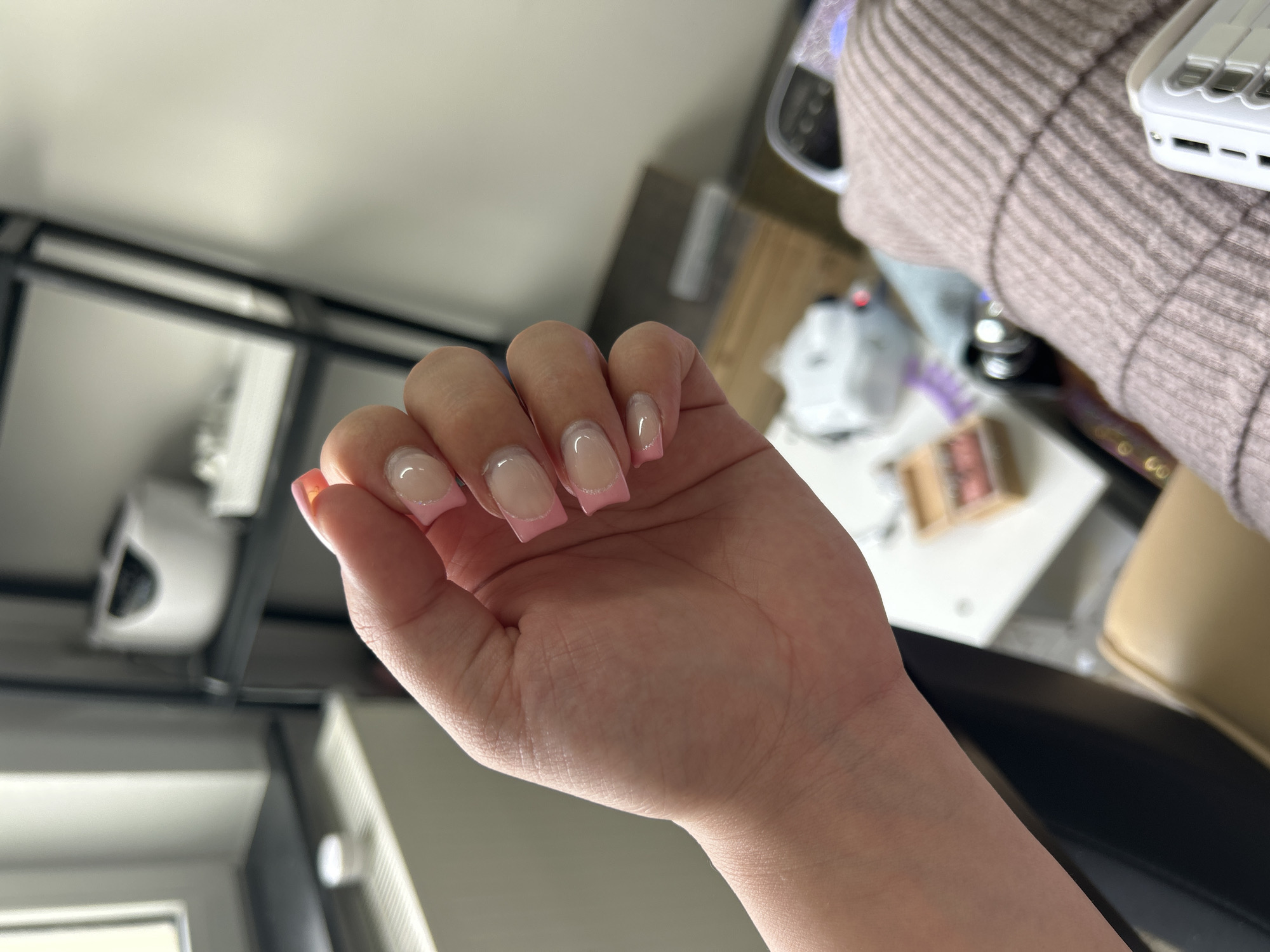 The image shows a close-up of a person's hand with freshly done, light pink and clear French tip acrylic nails. The hand is positioned in the foreground, obscuring much of the background. In the blurred background, several items are visible on shelves and a table. These include what appears to be a nail lamp, nail polish bottles, and other salon equipment. The setting seems to be a nail salon or a home-based nail studio. The image does not provide any specific information or visual cues that would allow for its location to be identified as Waldbröl, Germany.