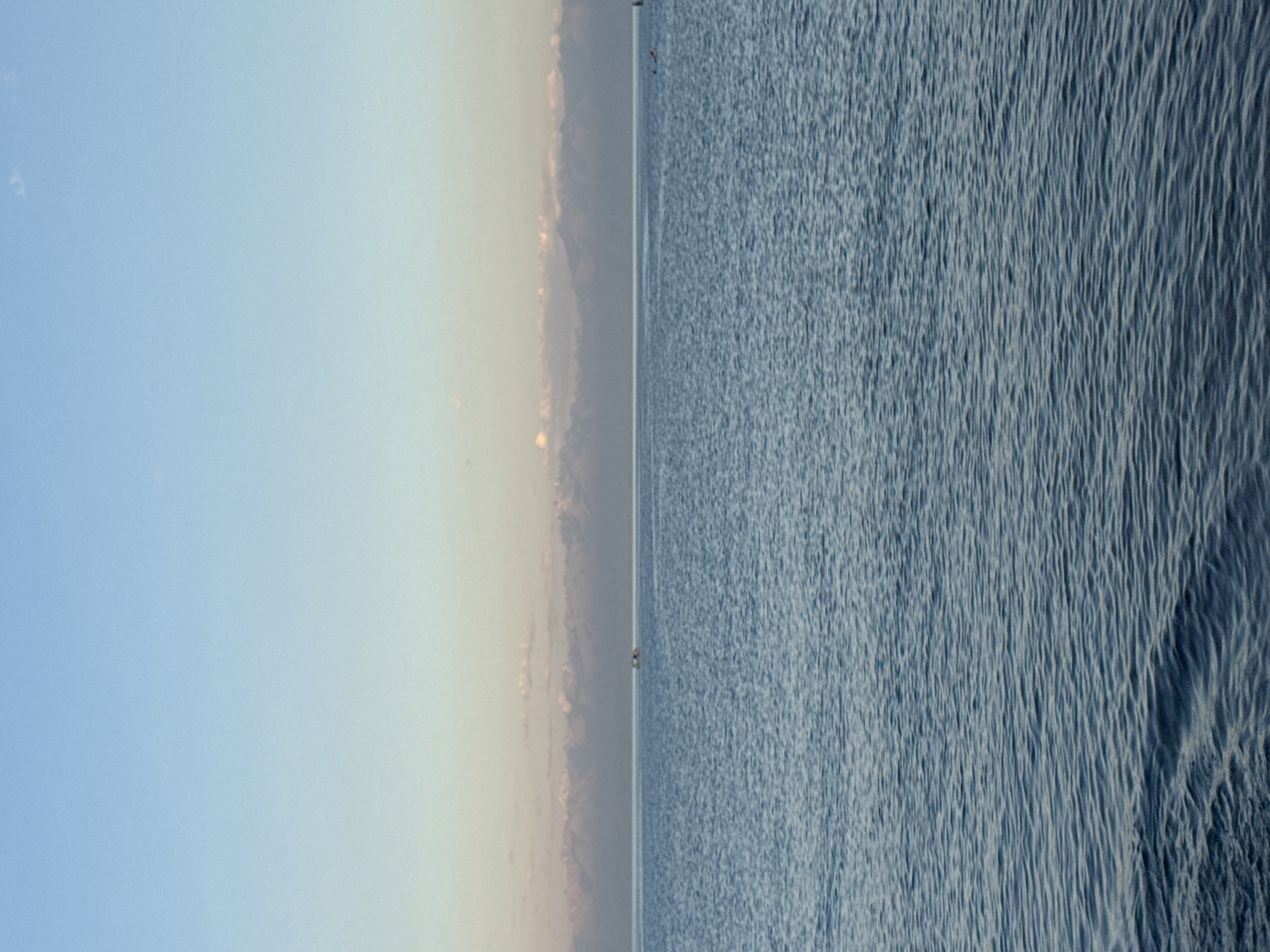 An expansive, calm body of water stretches across the foreground of the image, reflecting the soft hues of the sky. The water exhibits subtle ripples and texture, indicating a gentle movement. In the mid-ground, a distant, hazy mountain range forms a horizontal line, its peaks softened by atmospheric perspective. Above the mountains, a thin band of pale clouds is visible against a gradient sky that transitions from a light blue at the top to a warmer, almost peach tone near the horizon. A small boat is visible on the water, a speck against the vastness of the scene. The overall impression is one of tranquility and natural beauty.