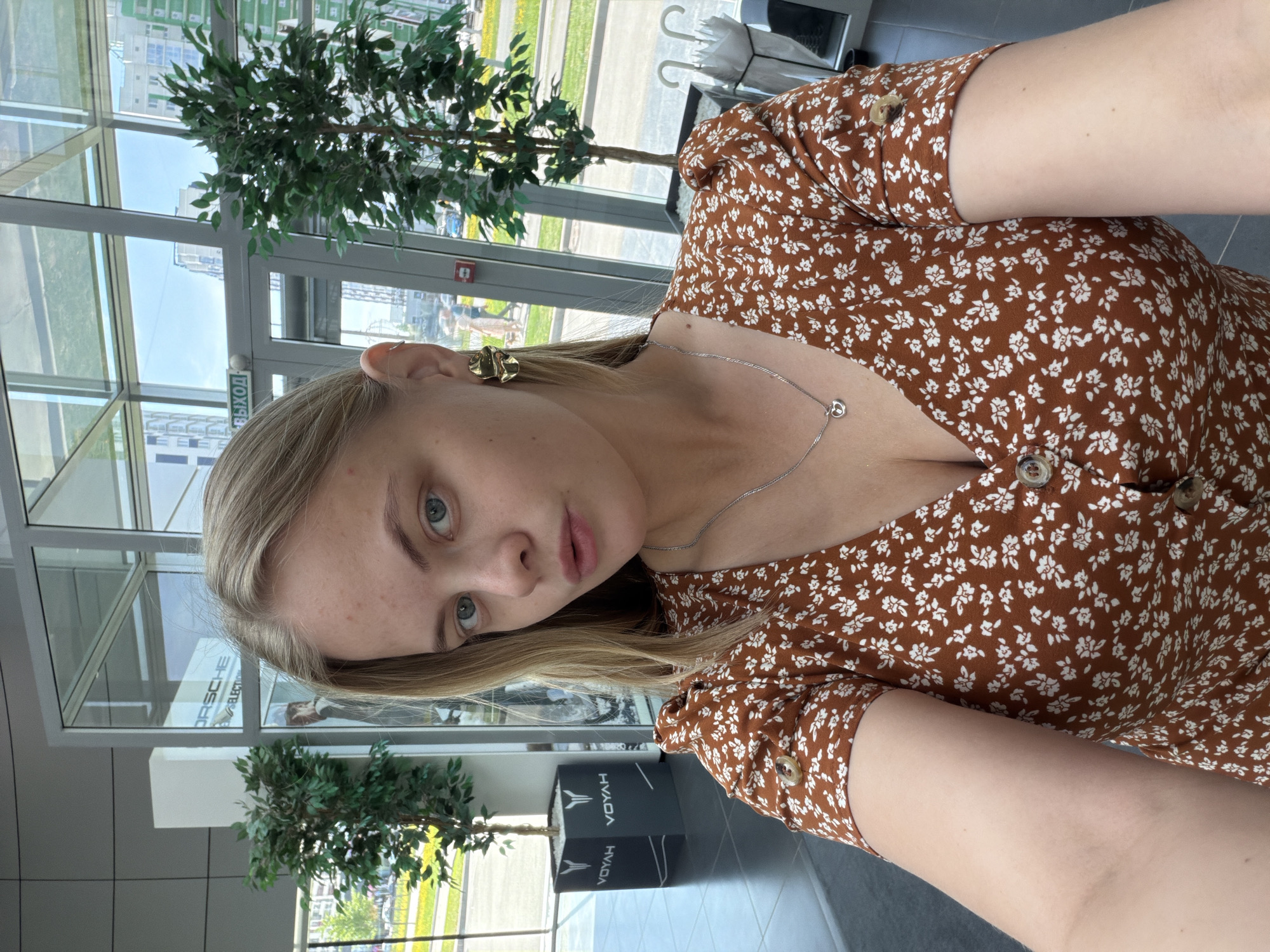 A woman with blonde hair and fair skin poses for a selfie indoors. She is wearing a brown, floral print dress and a silver necklace. Visible on her left ear is a gold earring. The background includes a commercial interior with large glass windows, a potted plant, and branding for Porsche and Voyah electric vehicles. The location is identified as Izhevsk, Russia.