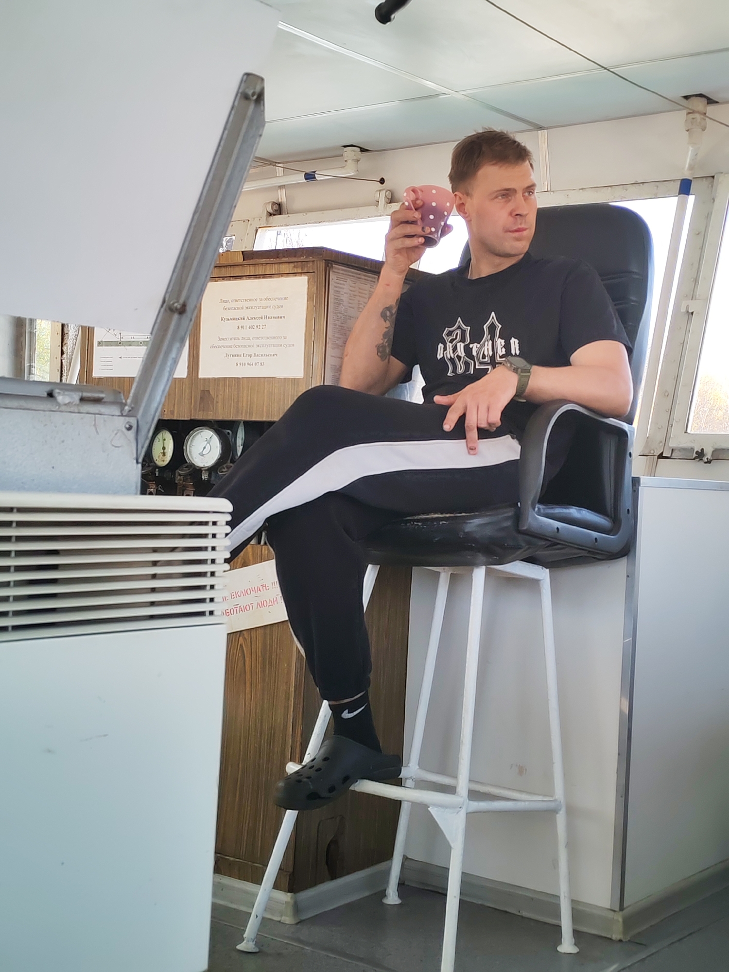 The image depicts a man sitting in a high chair inside what appears to be the control room of a vessel. He is holding a polka-dotted mug to his lips, wearing a black t-shirt with a graphic design, black athletic pants with a white stripe, black socks with the Nike logo, and black clog-style shoes. He has short, light brown hair and is glancing to the right. His right arm has a tattoo.
The man is seated next to a mechanical control panel and a wooden cabinet with gauges. A sign written in Russian is affixed to the cabinet, with text including phone numbers. The background shows a window with a view of the outside.
Based on the sign in Russian, the location might be related to the safety and operation of vessels. The OCR mentions phone numbers and names, indicating it could be a contact list related to vessel operation. Without further context, the precise location in the city of Dmitrov, Russia, cannot be determined.