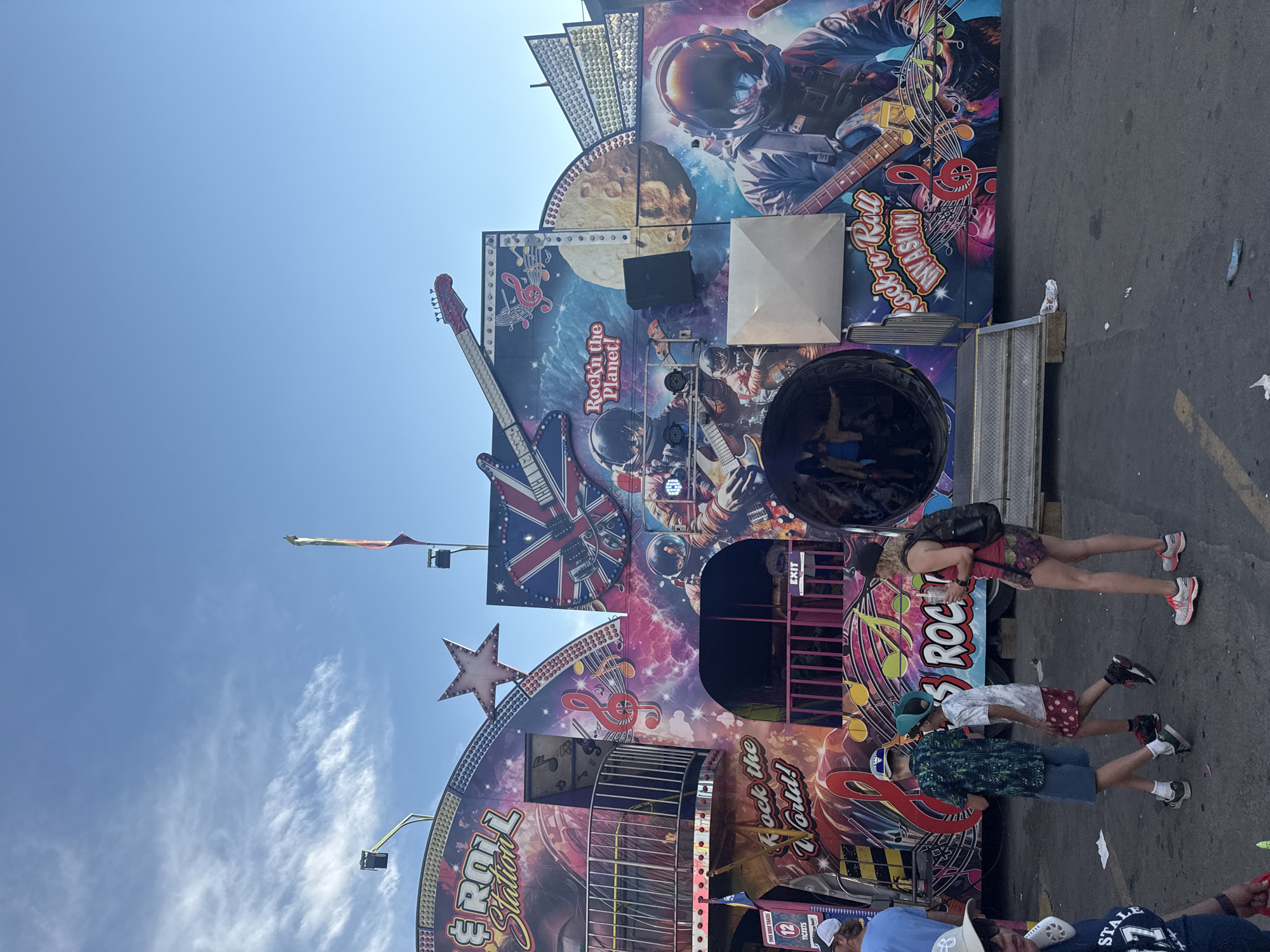 The image captures a lively scene at a carnival or fair, with a prominent ride adorned with a vibrant "Rock 'n' Roll Invasion" theme. The ride features large graphics of astronauts playing guitars against a backdrop of stars and planets, along with a stylized Union Jack flag. Text on the ride includes "Rock the Planet!" and "Rock 'n' Roll Station". Several people are visible, including children walking past the ride and individuals standing near an entrance. The environment suggests an outdoor event with asphalt ground and a blue sky with scattered clouds. The presence of a ticket booth with the number "12" and "Tickets" printed on it, along with what appears to be a ride exit, indicates this is an operational amusement attraction. The specific location in Calgary, Canada cannot be determined from the visual information provided in the image.