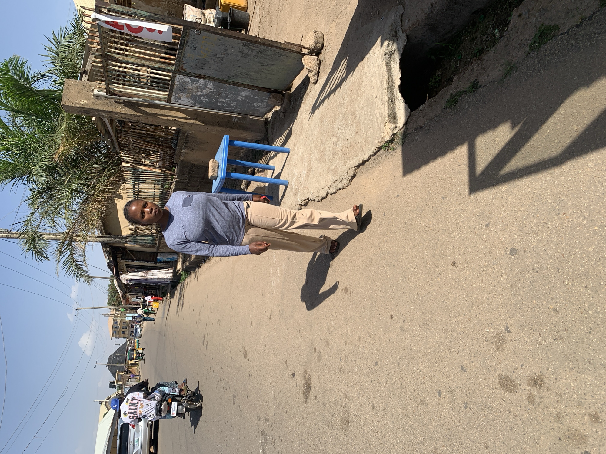 A medium-shot depicts a person standing on a paved street in Jos, Nigeria. The individual is facing forward and dressed in a long-sleeved blue shirt and tan pants. They are standing next to a blue plastic chair and a concrete sidewalk bordering a building with a gate and foliage on the left. The street is dusty, and in the background, a motorcycle with two people on it is moving away. A drainage ditch is visible on the right of the frame, casting a shadow on the street. Other vehicles and people are visible further down the street. The sky is blue and hazy.