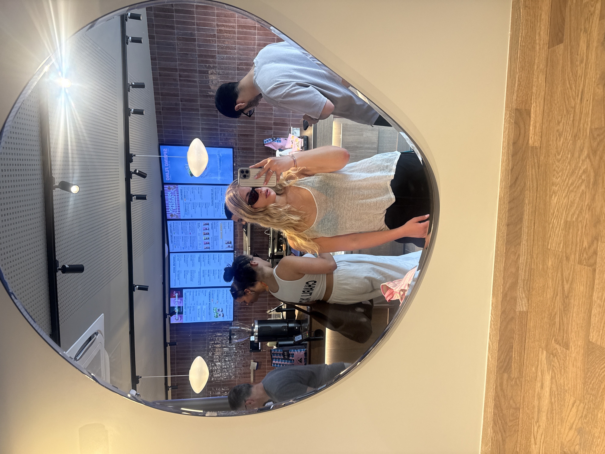 Captured in a reflective oval mirror, the image presents a scene from what appears to be a café or eatery. Reflected in the mirror is a woman taking a selfie with her smartphone. Her blonde hair is styled in loose waves, and she wears sunglasses. A man with a beanie is visible next to her, seemingly looking at something on a counter. Further back, another woman with a bun and wearing a tank top is partially visible, as is another man with a light-colored shirt.
The mirror reflects the interior of the establishment, revealing menu boards with text, hanging lights, brick walls, and other customers. This likely indicates that the location is a restaurant or café, possibly in Paris, France.
The image primarily captures the actions of individuals taking selfies and interacting within the café environment. The mirror acts as a frame, capturing and reflecting a slice of the café's interior and the activities within.