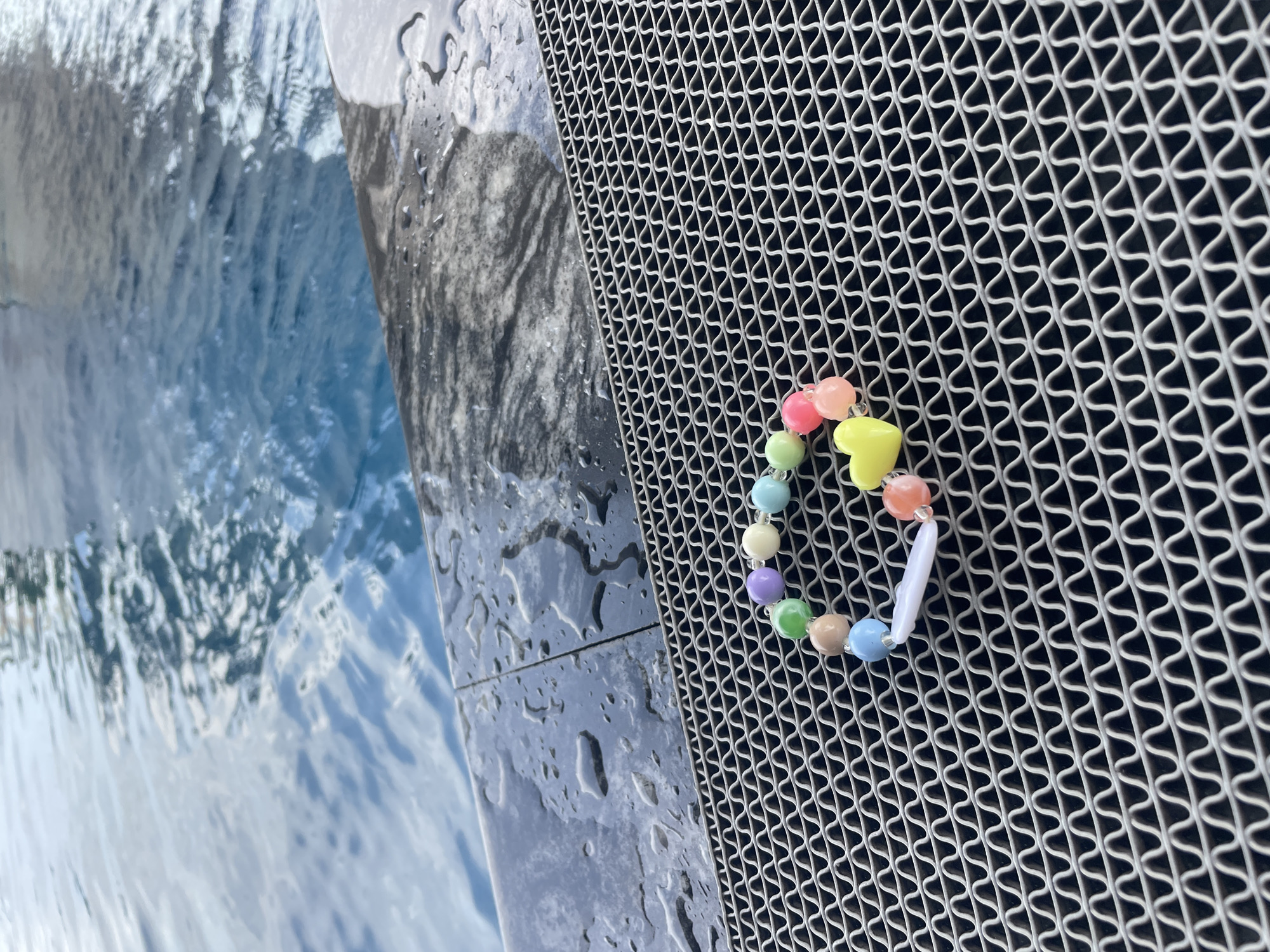 A colorful beaded bracelet lies on a gray ribbed mat, positioned at the edge of a swimming pool. The bracelet features beads in pastel shades of purple, light blue, green, and pink, along with a yellow heart-shaped charm and a white oval charm. The pool water is a vibrant blue, with ripples and reflections suggesting movement. The edge of the pool is made of dark gray stone with visible water droplets. The scene is outdoors, and the lighting indicates it is daytime. The location is identified as Nha Trang, Vietnam.