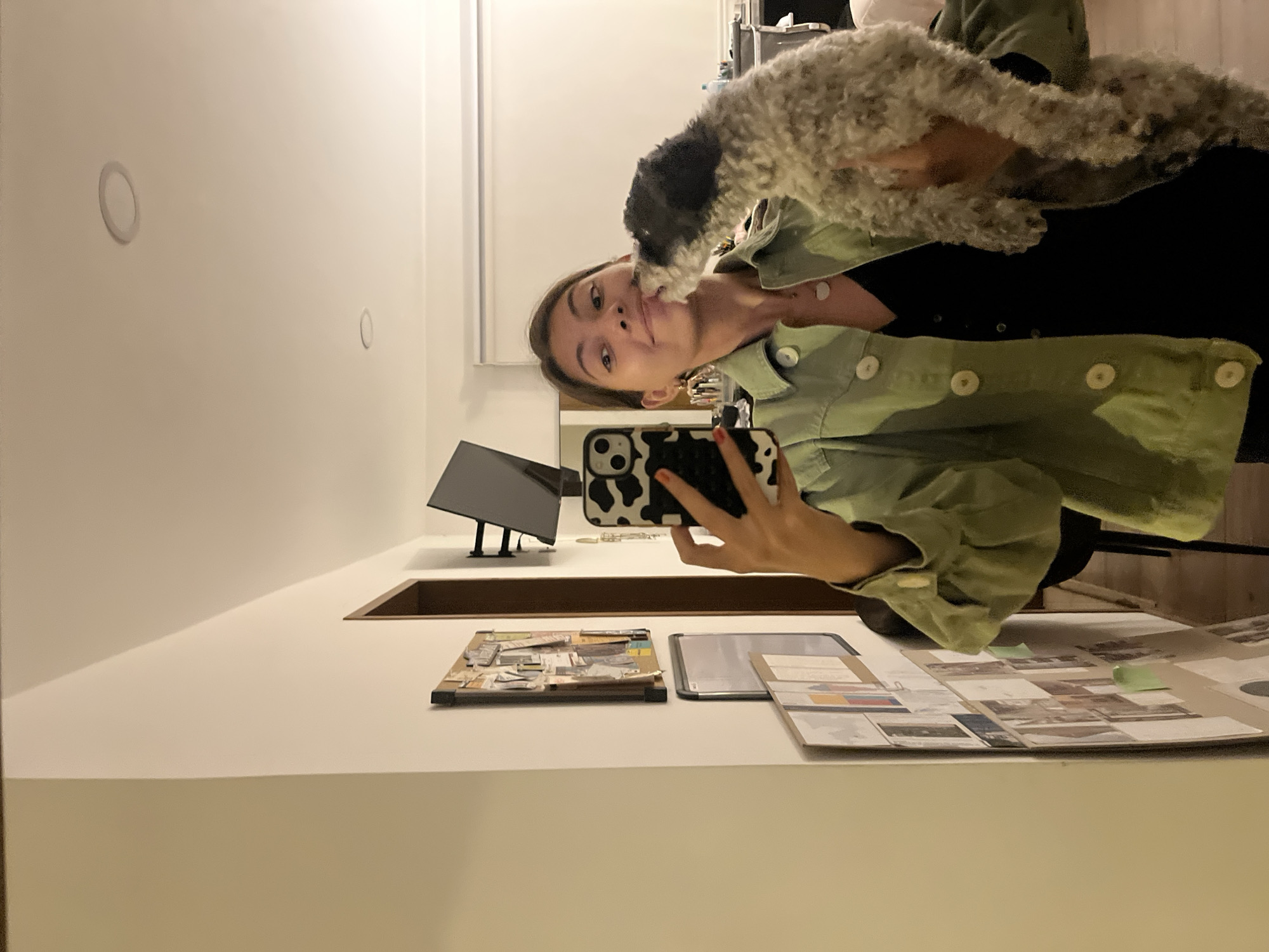 A woman is taking a selfie with a gray and black dog indoors. The woman is wearing a green button-down shirt over a black top and holding a cell phone with a black and white cow-print case. A desk or counter in front of her holds a cork board with images, a gray tablet, and a spread of photographs or design materials, some with green sticky notes. A computer monitor is visible in the background. The location is not discernible from the image.