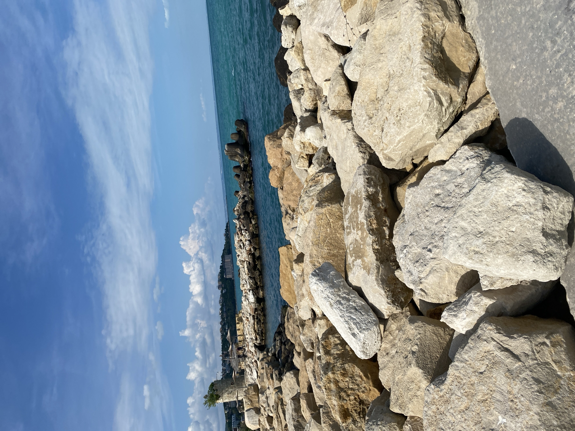 The image depicts a rocky shoreline in an urban setting. Large, light-colored rocks dominate the foreground, forming a barrier against the turquoise water. A city skyline is visible in the background, suggesting a coastal urban environment. The sky is blue with scattered clouds. The location is Unknown, Unknown.