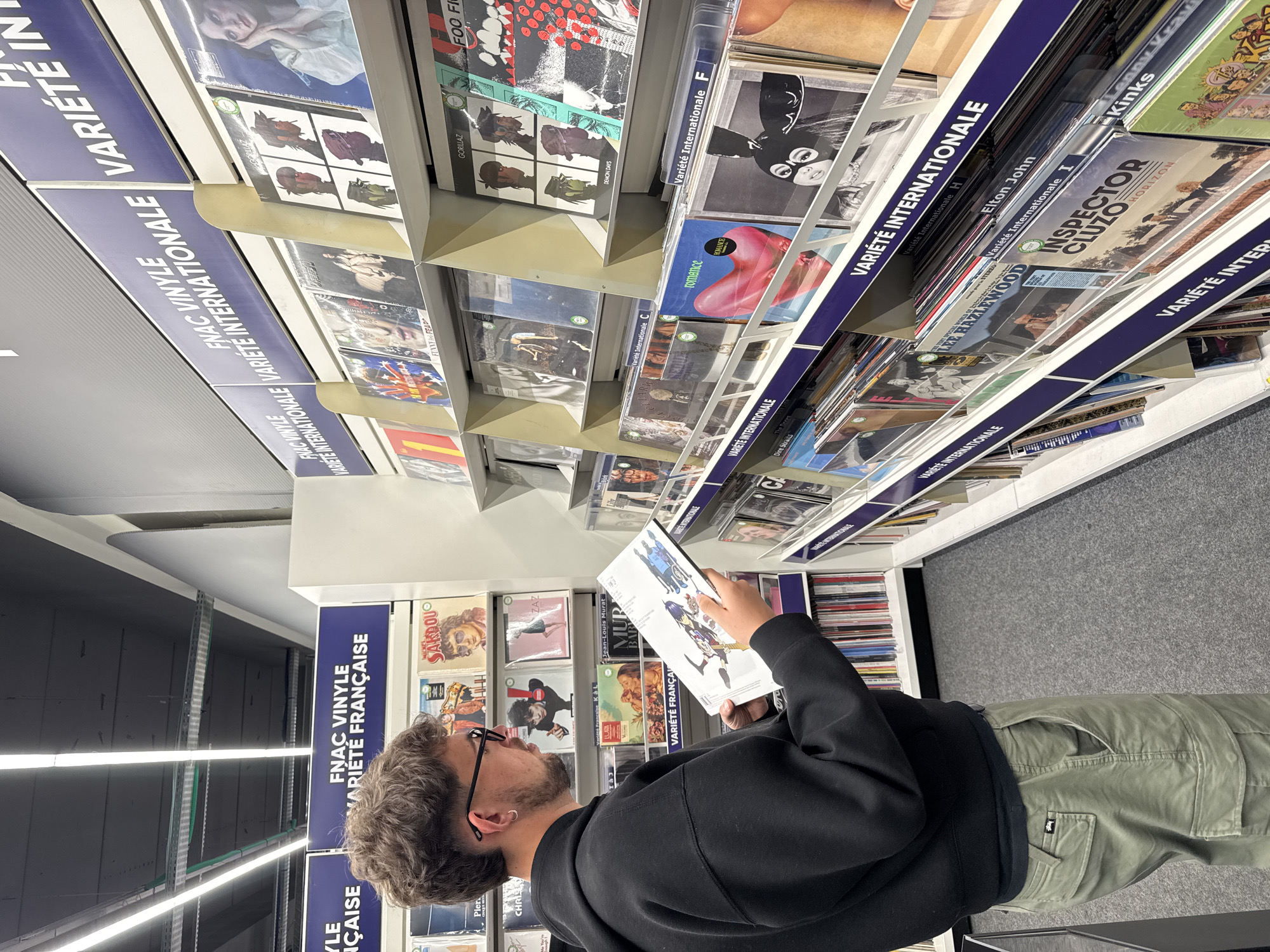 A man with curly hair, wearing glasses, a black hoodie, and green cargo pants, is looking at a vinyl record in a store. He is standing in the "FNAC VINYLE VARIÉTÉ FRANÇAISE" section, surrounded by shelves filled with vinyl records, categorized by sections such as "Variété Internationale". The labels are in French. The location is possibly a FNAC store.