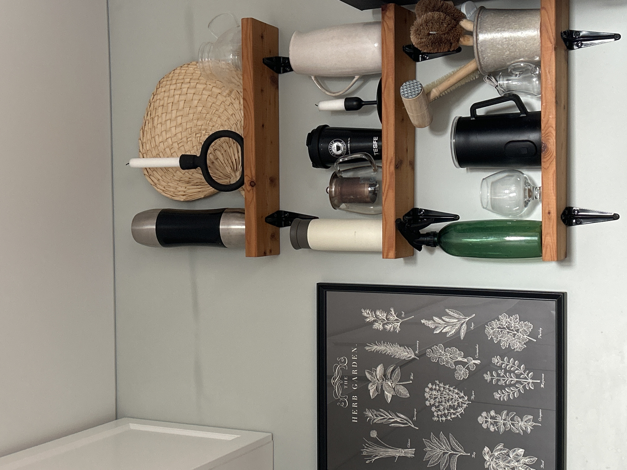 A vertical photograph shows a wooden rack holding various kitchen and bar items. The rack is mounted on a pale green wall. On the left side of the rack, a straw-like woven disk is displayed. Below it, a black and silver insulated tumbler is stored horizontally.
The rack itself is divided into sections. In the upper left section, a white mug is placed. To its right, a smaller white object with a black handle is positioned horizontally. Next to this, a black cylindrical device with a logo that reads "TESFE" is visible. Below the mug, a clear glass with a handle rests on its side.
Further to the right on the rack, a wooden-handled brush with brown bristles is at the top. Below it, a silver-toned metal container is situated. A small glass bottle with a handle is placed below the silver container. Another black cylindrical device with a handle is in the middle section of the rack. Below this, a wine glass is held horizontally. Finally, a long, green glass bottle occupies the bottom section of the rack.
Below the wooden rack, a framed black and white print of botanical illustrations of herbs is visible. The text "THE HERB GARDEN" is prominent on the print. The overall scene suggests a kitchen or pantry area, organized and somewhat decorative. No people are present, and the lighting suggests an indoor setting during the daytime.