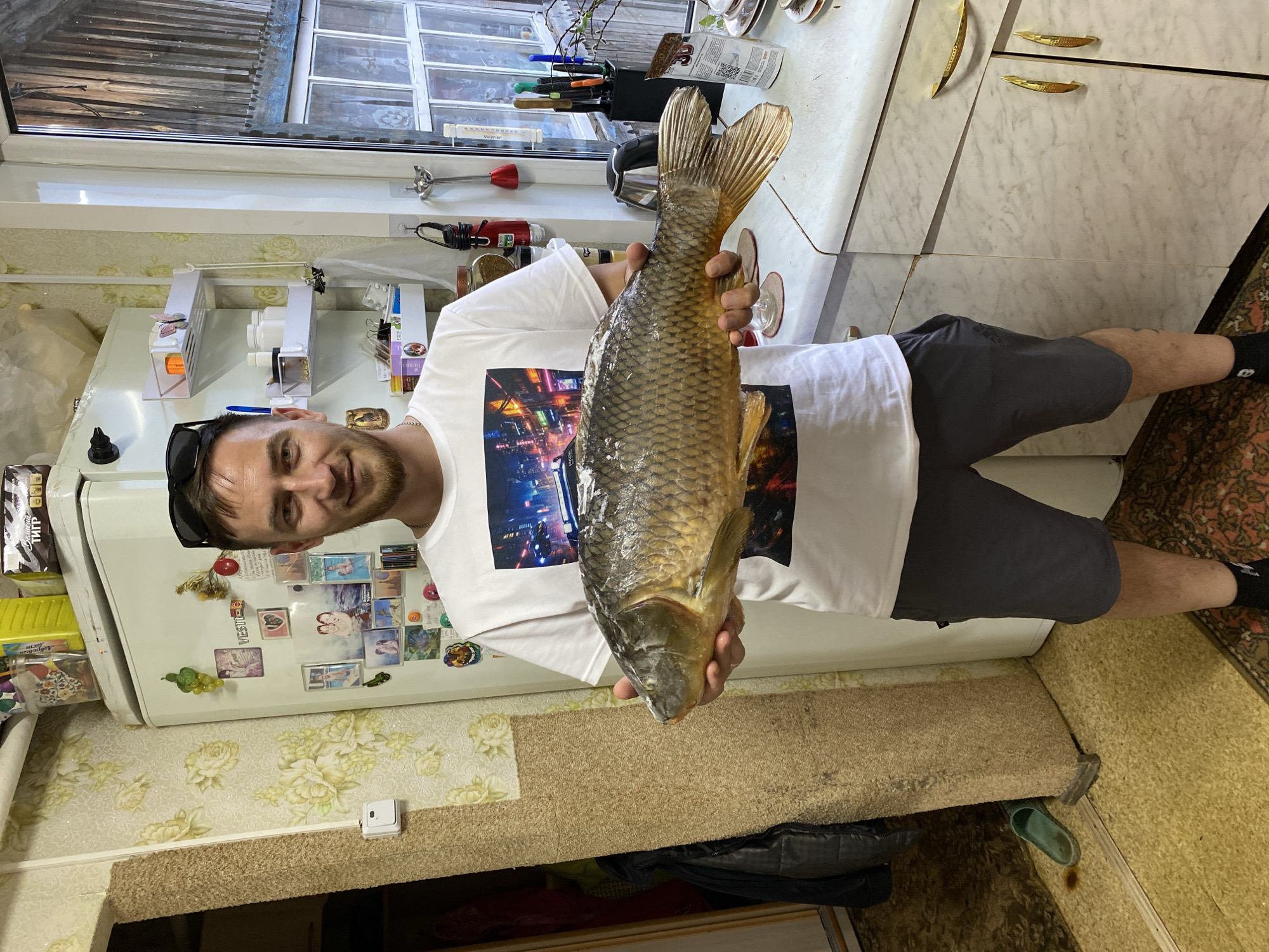 A man is holding a large fish in a kitchen. The man is wearing shorts and a t-shirt with a design. A refrigerator with various items on it is visible in the background, along with a window and kitchen cabinets. The floor is carpeted. The location is Баклуши, Russia.