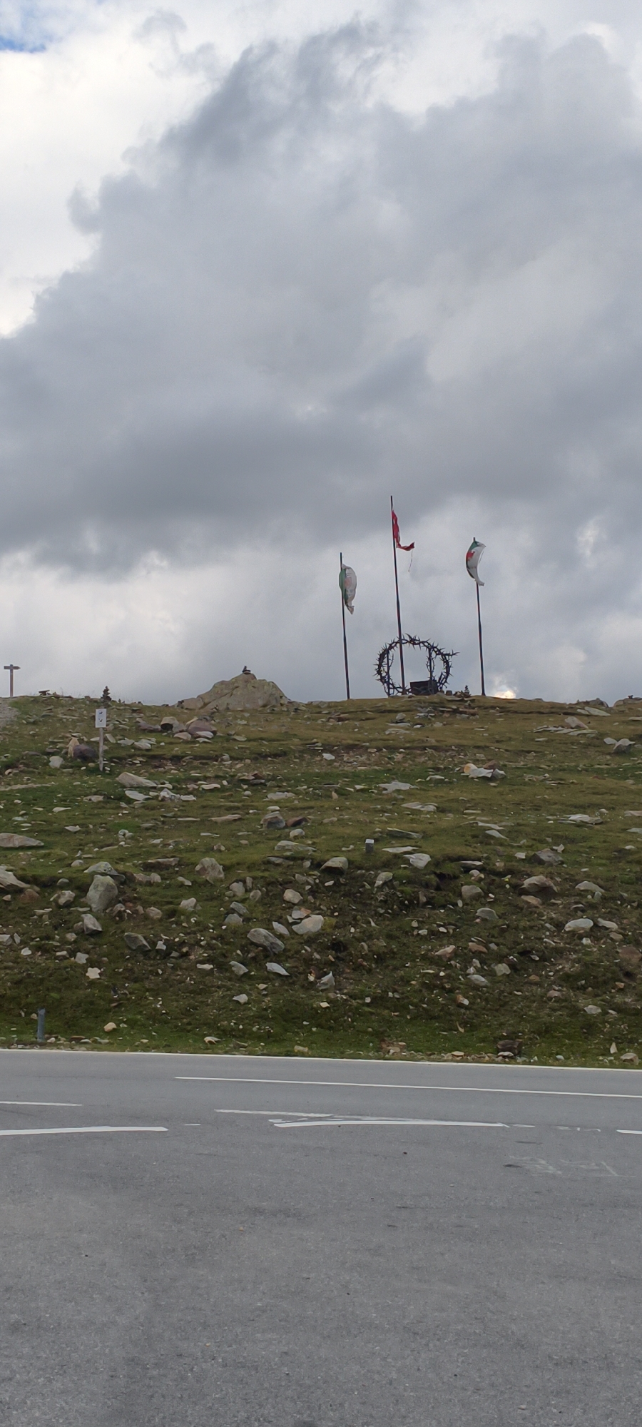 The image shows a view from the side of a road, looking up at a grassy, rocky hillside. In the distance, atop the hill, are three flagpoles with flags, and a circular metal structure with sharp spikes resembling a crown of thorns. There are also several cairns (piles of stones) on the hill. The sky is overcast with dark clouds. The road in the foreground has white lane markings. There are no people visible in the image. The scene appears to be in a mountainous, possibly alpine, region.