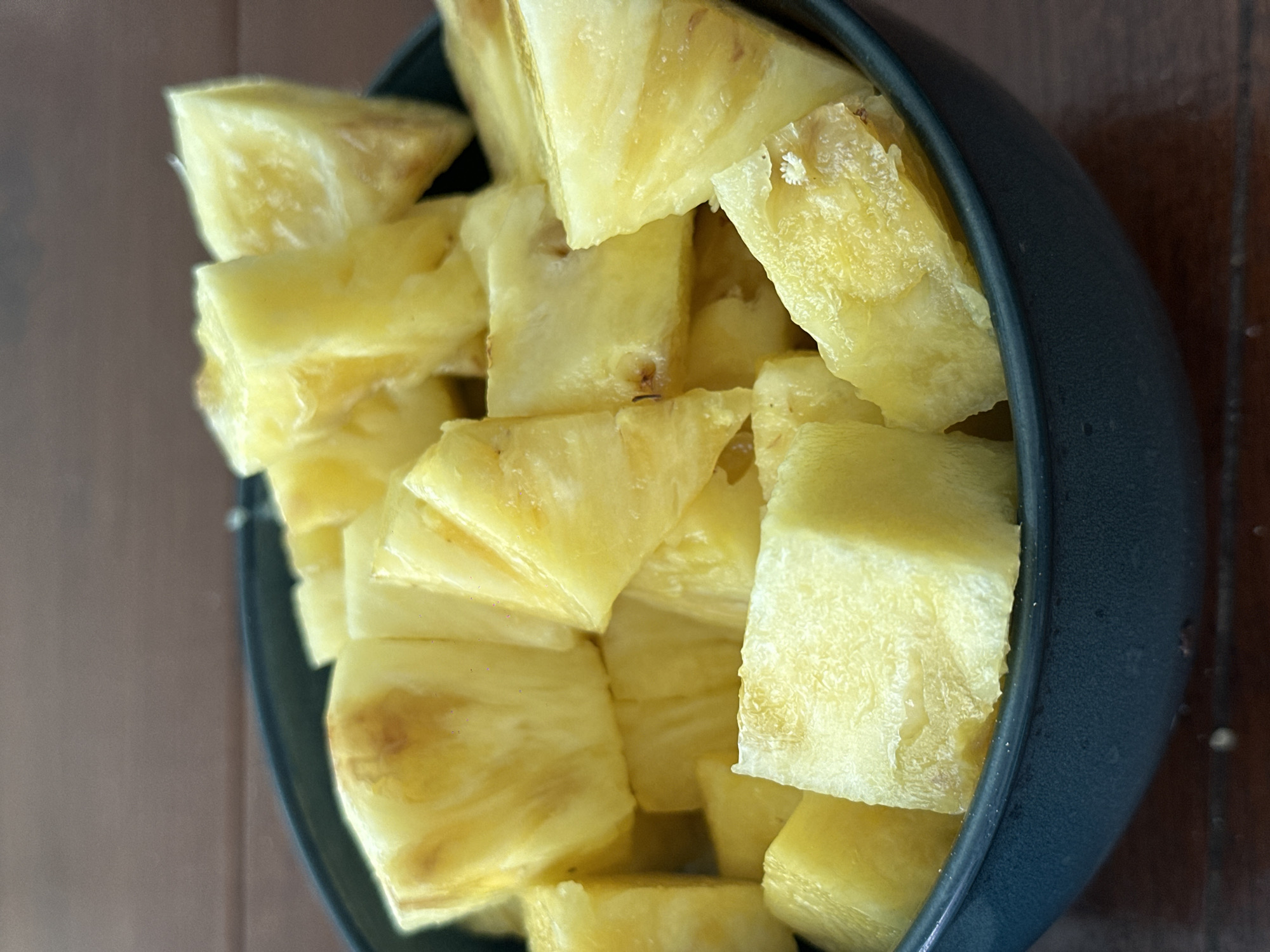 A close-up shot shows a bowl filled with chunks of pineapple. The bowl is dark gray and sits on a wooden surface with a visible grain pattern. The pineapple pieces are yellow and appear freshly cut. No people or any indication of the location in Amsterdam, Netherlands, are visible in the image.