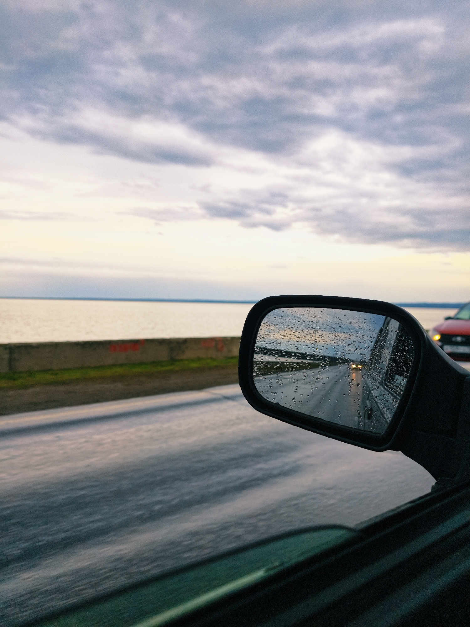 A photograph taken from inside a moving vehicle shows a view of the road and the surrounding environment, partially captured in the side mirror. The road is wet, reflecting the overcast sky. The sky is filled with gray and white clouds. To the left, a body of water is visible behind a low concrete barrier with patches of green vegetation. The side mirror of the vehicle is covered in raindrops and reflects the road behind the vehicle, with a car's headlights visible in the distance. A red car is partially visible behind the side mirror. The inside of the vehicle's door is visible in the lower part of the frame. The location is Chaikovsky, Russia.