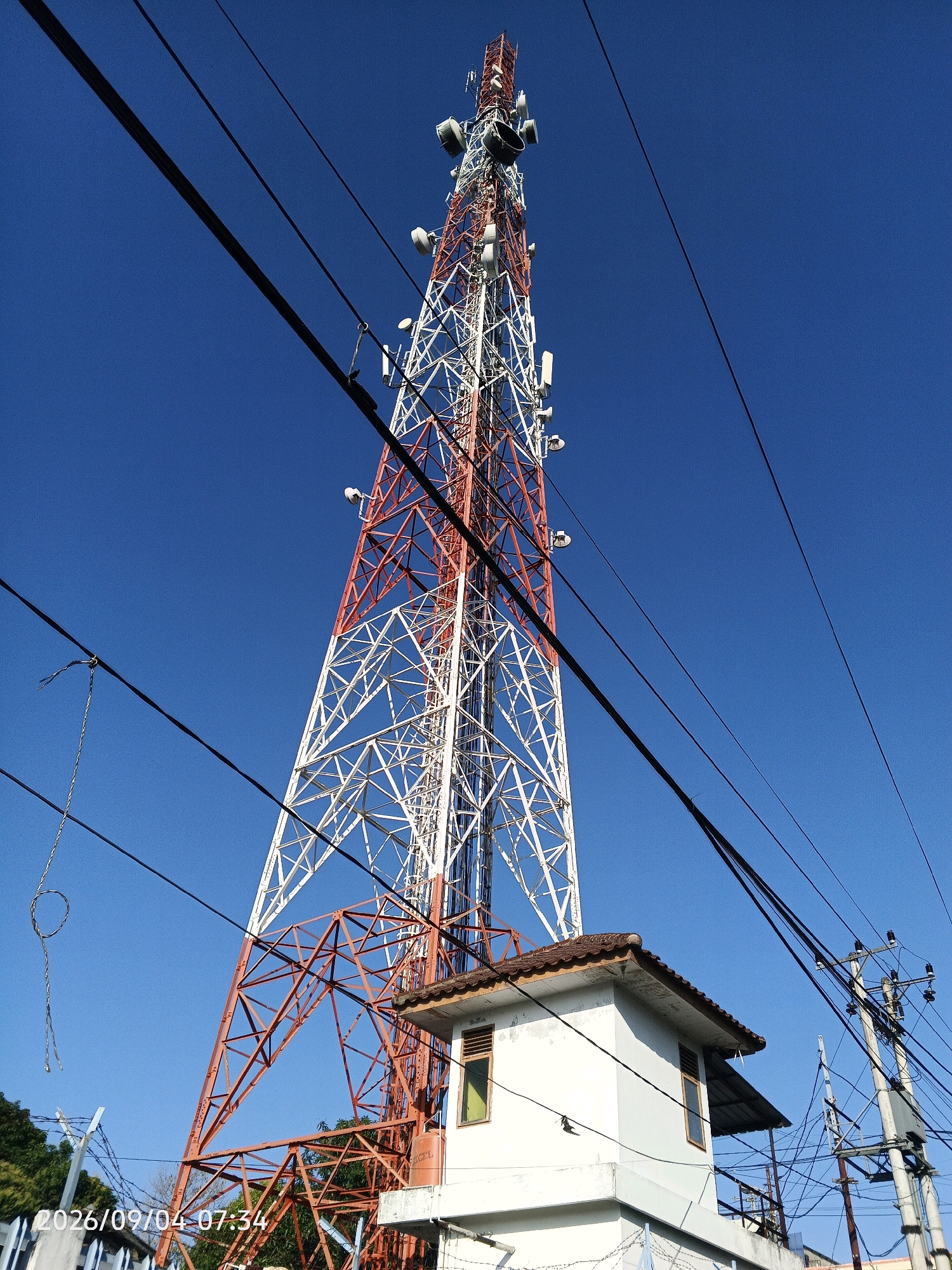 A low-angle, wide shot captures a tall telecommunications tower against a clear blue sky. The tower, painted white and red, features numerous antennae and communication equipment. In the foreground, a small, white building with a tiled roof sits to the right, with power lines crisscrossing the frame. The date stamp "2026/09/04 07:34" is visible in the bottom left corner. The scene appears to be in an urban or semi-urban environment in Pare-Pare, Indonesia, given the context of the date stamp. There are no people visible in the image.