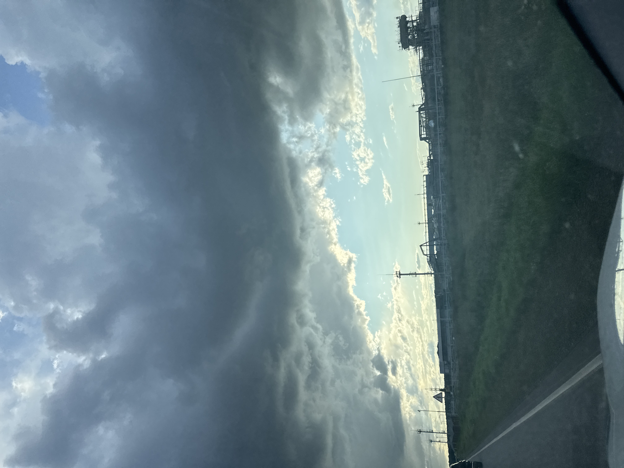 The image depicts an industrial landscape with a cloudy sky, likely taken from a moving vehicle on a road. In the foreground, a portion of a road with a white line is visible, along with the edge of a vehicle's dashboard. The middle ground features a grassy field stretching towards a line of industrial structures. These structures appear to be part of a factory or processing plant, with various pipes, towers, and buildings. Several tall poles with antennas or lights are also present. The background is dominated by a dramatic sky filled with dark, heavy clouds, with patches of lighter, brighter sky and some scattered white clouds visible. The scene suggests an overcast or impending stormy weather condition. The location is identified as Ptitsefabrika, Russia.