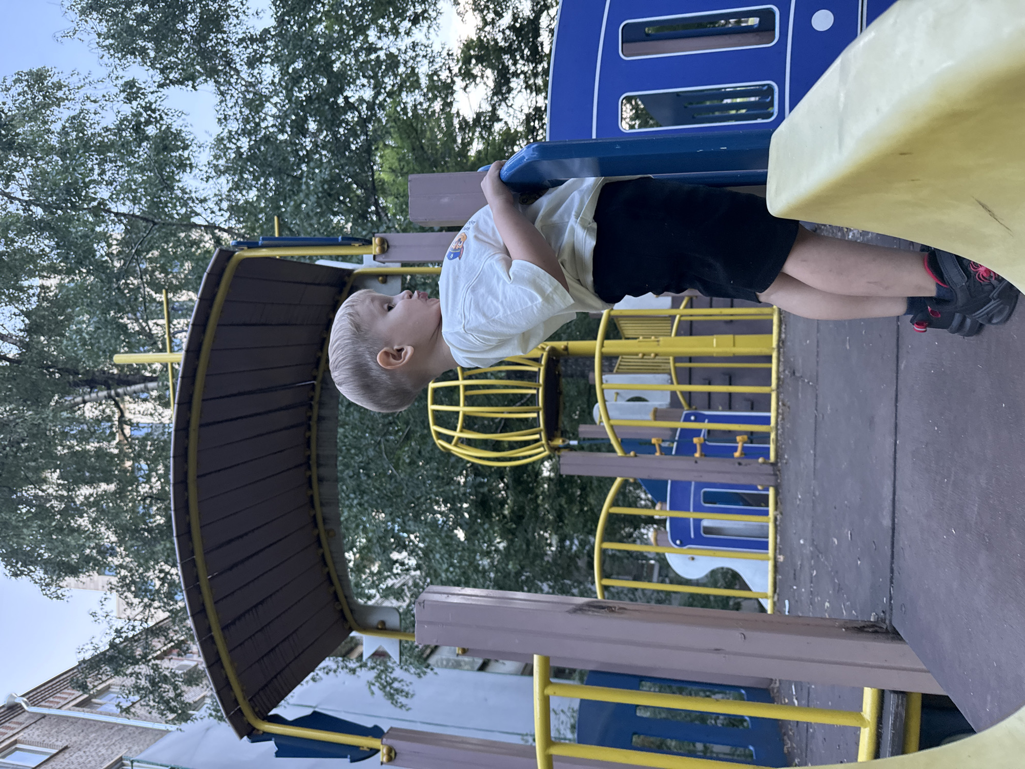 The image shows a young boy on a playground. He is wearing a white t-shirt and black shorts. He is standing on a wooden platform with a blue railing. The playground equipment is colorful, with yellow and blue accents. In the background, there are trees and a building. The image appears to have been taken in Moscow, Russia, as indicated by the architectural style of the building and the type of playground equipment. The boy is looking off to the side, and his mouth is slightly open, as if he is about to speak or is surprised by something. The overall mood of the image is one of childhood innocence and playfulness.