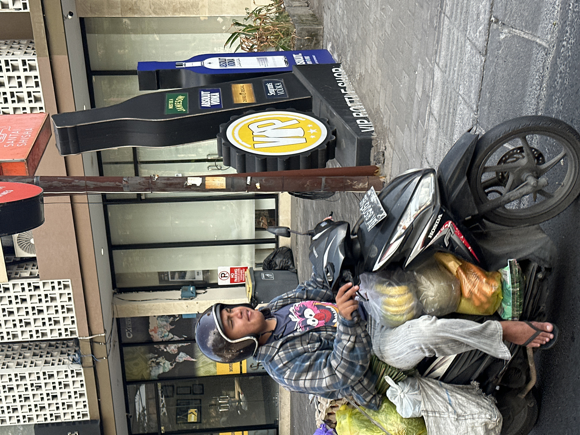 A man rides a motorcycle through Denpasar, Indonesia, carrying a load of produce. He is wearing a helmet and a plaid shirt, and is riding on the right side of the road. The background shows a building with a sign that reads "VIP BOTTLE SHOP". The street is paved with concrete, and there are trees and other vegetation on the side of the road. The sky is overcast.