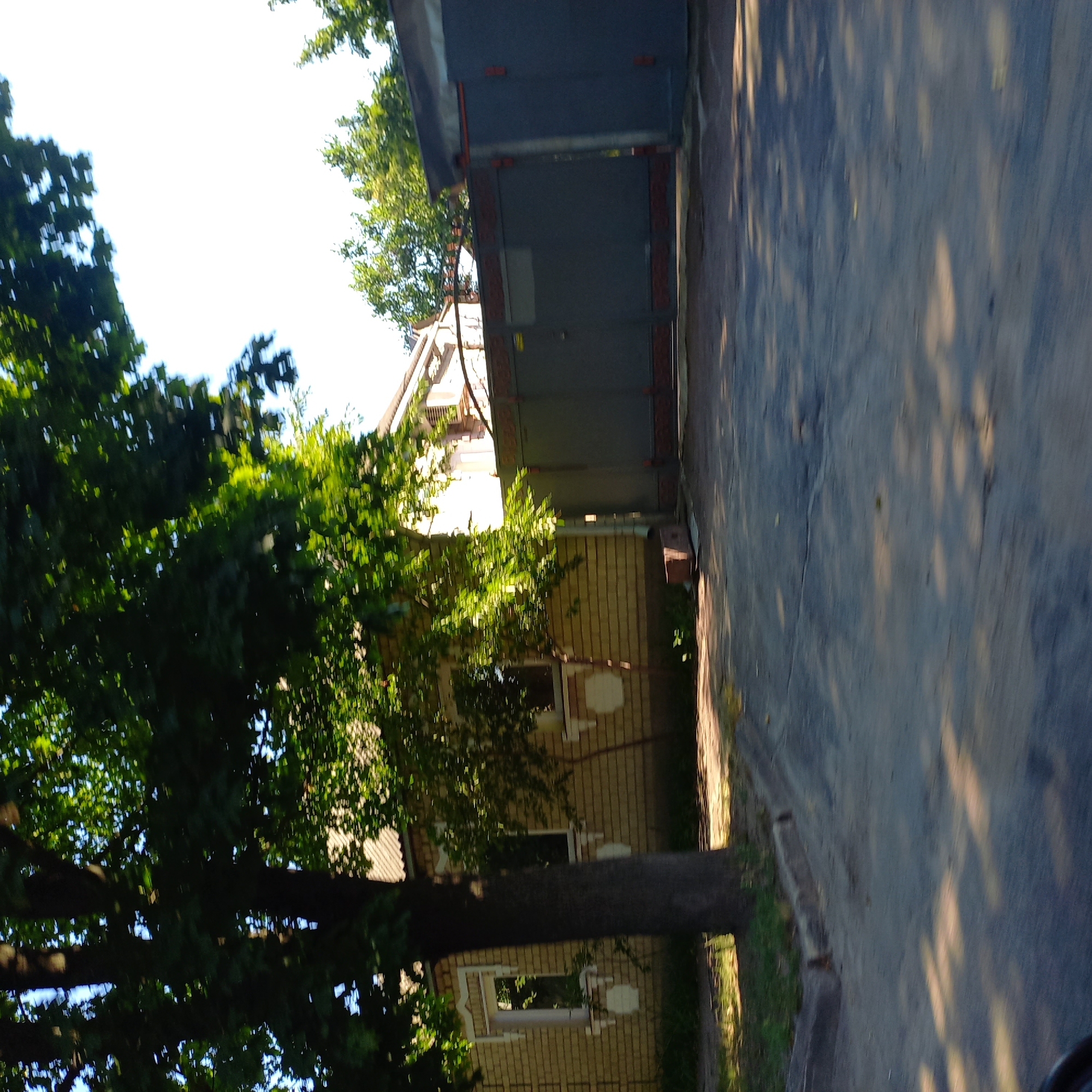 The image captures a view from a street in Kropyvnytskyi, Ukraine, showing a brick building with a metal gate and a large tree in the foreground. The building, constructed with light-colored bricks, has a roof and visible windows with white trim. In front of the building is a dark gray metal gate, partially obscured by the foliage of a large tree. The tree, with its dark trunk and green leaves, dominates the left side of the frame, casting shadows on the paved surface of the street. The street itself appears to be asphalt, with some visible cracks and debris. The lighting suggests it is daytime, with sunlight filtering through the leaves, creating dappled patterns on the ground. The overall scene depicts a residential or commercial property in a Ukrainian urban setting.