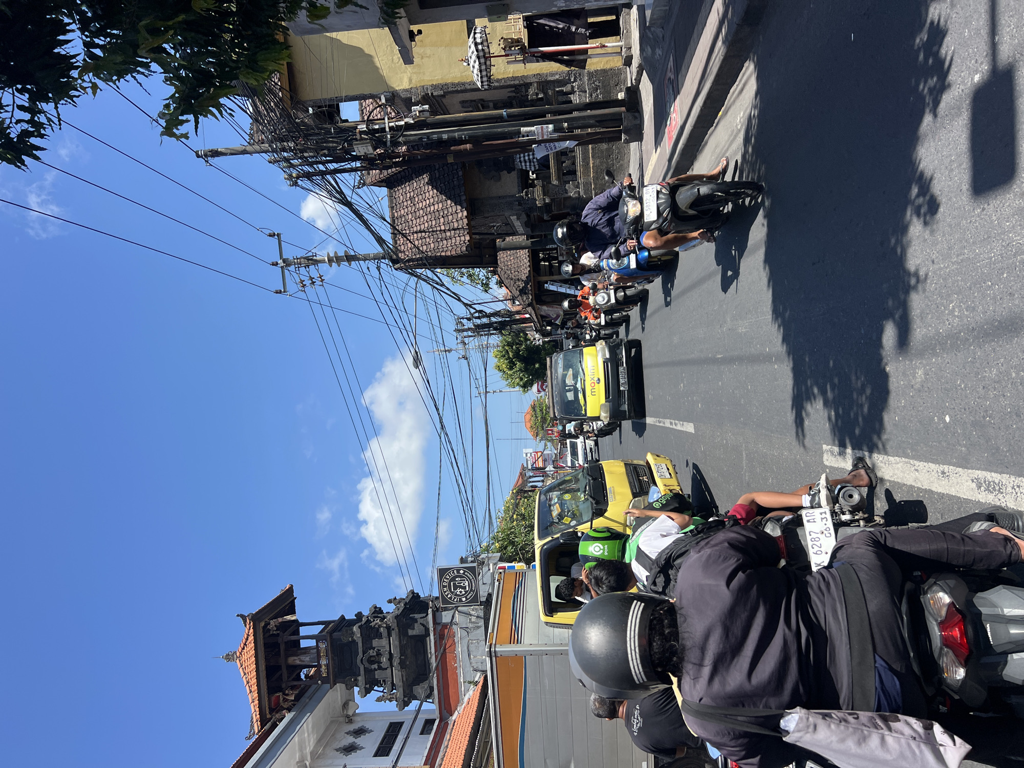 The image depicts a busy street scene in Denpasar, Indonesia, likely on a sunny day with a clear blue sky. The foreground is dominated by a procession of motorcycles and a yellow truck, indicating traffic congestion. Several individuals are visible on motorcycles, many wearing helmets. The architecture in the background suggests a tropical urban environment, with traditional Balinese structures featuring tiered roofs and intricate stone carvings, such as the Pura Dalem Sakenan tower visible on the left. Overhead, a dense network of electrical wires crisscrosses the sky, supported by utility poles. The road surface is asphalt, and the sides of the street are lined with buildings, some appearing older with terracotta roof tiles and others more modern. A sign for "Flash Device" is affixed to a building. The presence of Gojek branding on a motorcycle helmet and the yellow truck suggests ride-sharing services are active. The overall impression is a vibrant, bustling city with a blend of traditional and modern elements, characteristic of Denpasar.