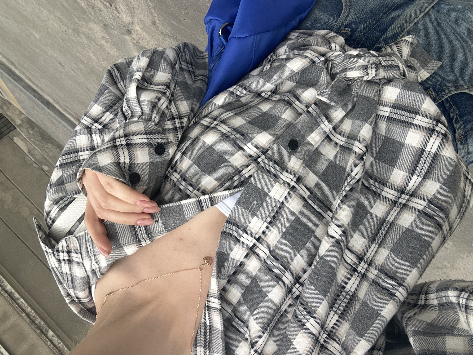 This close-up, high-angle photograph captures a partial view of a female subject outdoors in Ternopil, Ukraine. The subject is wearing a grey, white, black, and faint beige plaid flannel shirt, unbuttoned to reveal a white undershirt underneath. A slender gold-toned chain necklace with an intricate, open-work heart-shaped pendant rests on her collarbone and upper chest. The subject's left hand, featuring manicured, almond-shaped fingernails painted a light pink or nude shade, rests gently on the front of the plaid shirt. On the lower right, distressed light blue denim jeans are visible. Adjacent to the jeans and the plaid shirt, a vibrant royal blue fabric bag, possibly a backpack or sling bag, is partially seen, showing a zipper and a silver-toned D-ring. The environment consists of a grey concrete ground surface, with textured concrete elements, possibly steps or a curb, visible in the upper left background. The lighting appears overcast, resulting in soft shadows.