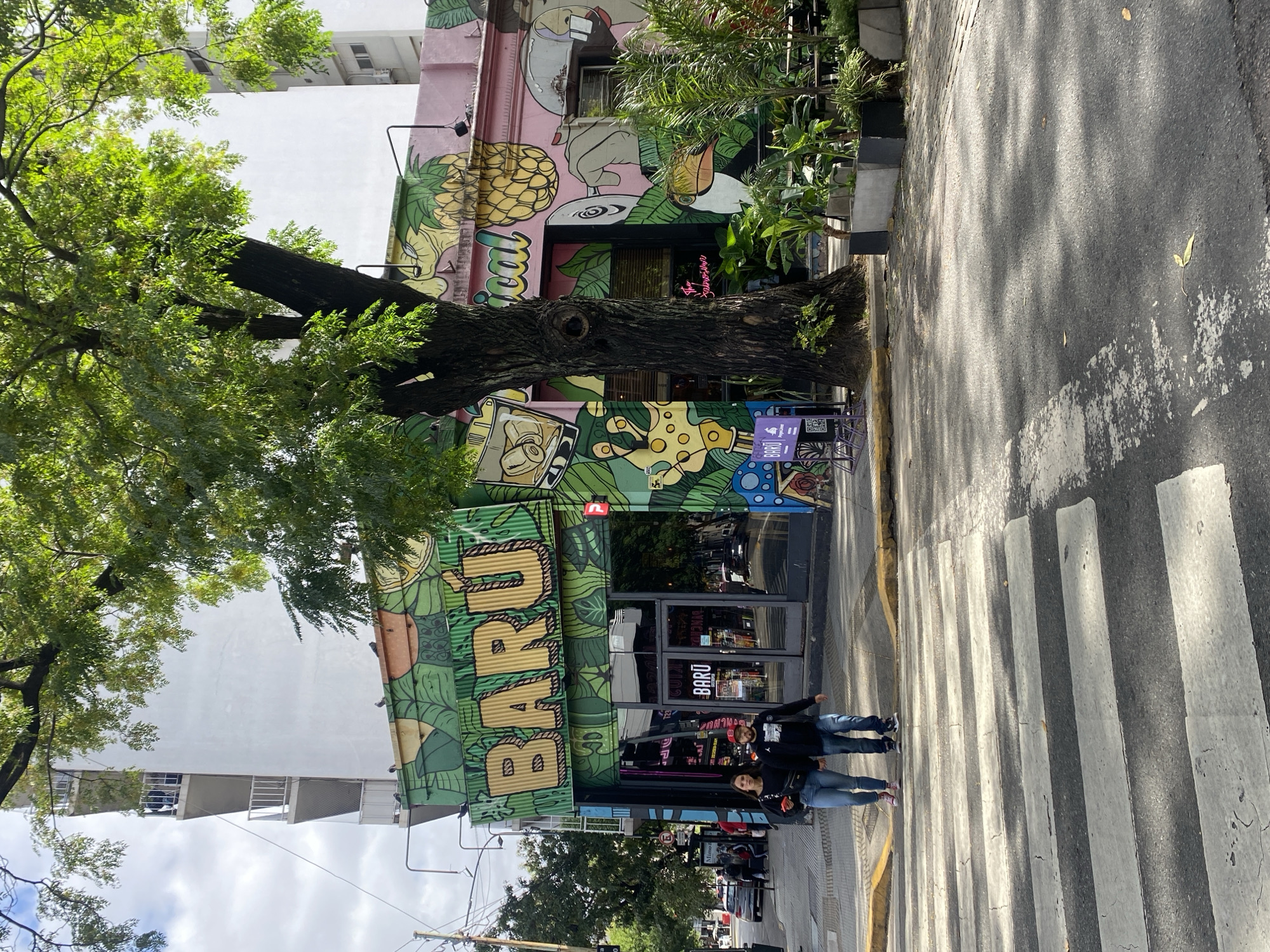The image shows a street scene in Buenos Aires, Argentina, featuring the "Barú" bar with colorful murals. The bar's facade is adorned with painted imagery including tropical fruits, cartoon-like figures, and the bar's name in large, bold letters. A mature tree stands adjacent to the building, its branches partially obscuring the bar's upper sections. Two individuals are standing on a crosswalk in front of the bar. The surroundings include a typical city street with buildings, a sidewalk, and other pedestrian traffic visible in the distance.