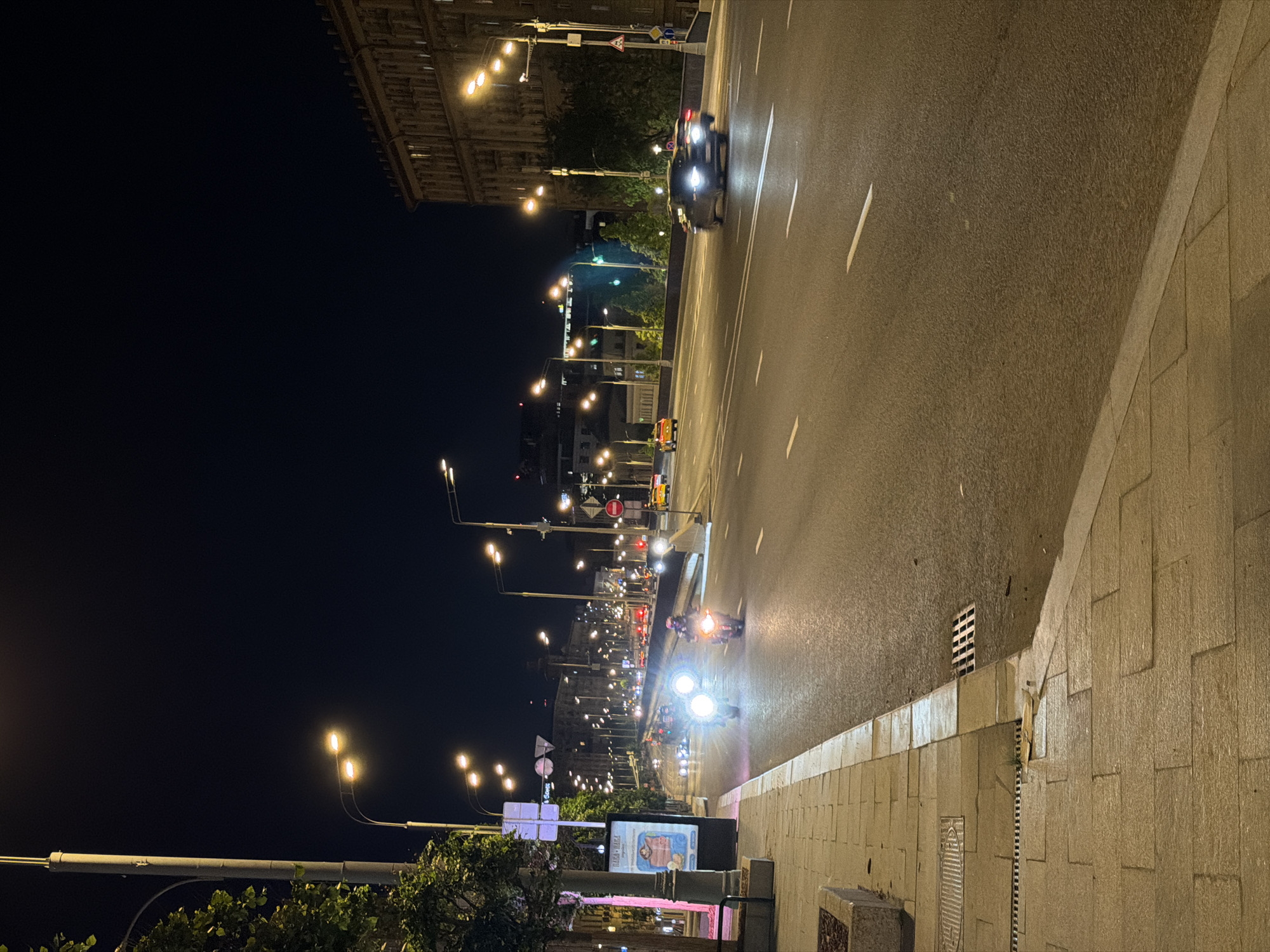 The image depicts a nighttime view of a street in Moscow, Russia. Several cars and motorcycles are traveling on the road. Streetlights illuminate the scene, and buildings are visible in the background. A sidewalk with paving stones runs alongside the road, and a billboard is present.