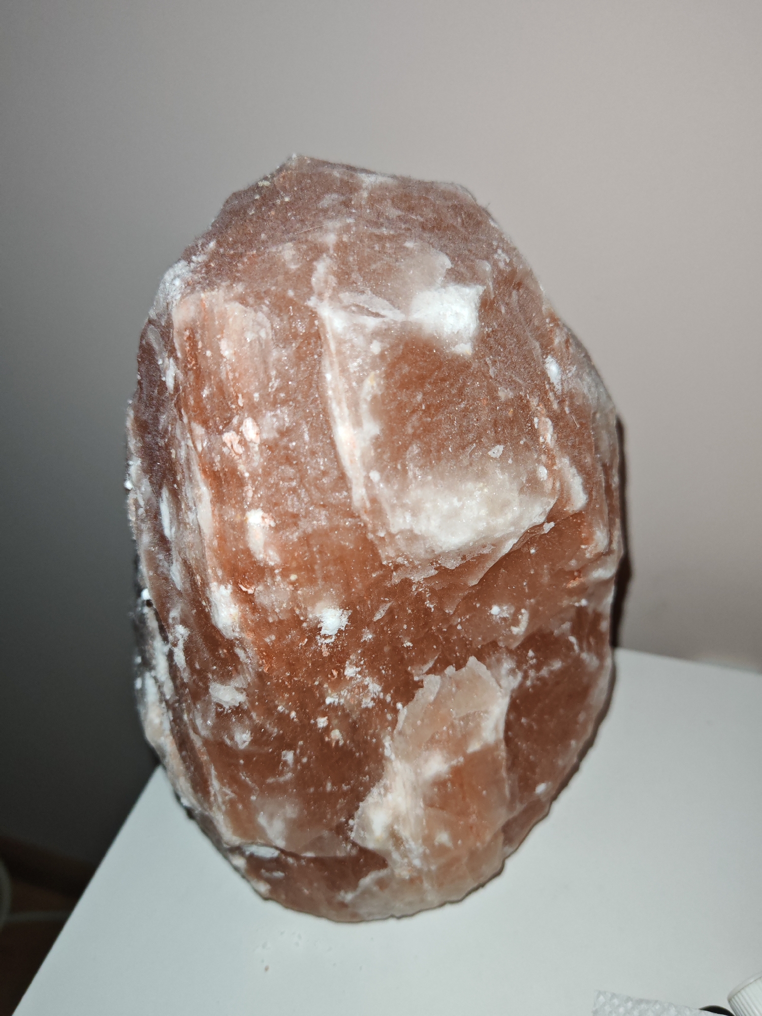 The image shows a close-up of a large, rough-hewn, pinkish-orange salt lamp. The surface of the lamp is irregular and appears crystalline, with white salt deposits visible in crevices and on some edges. The lamp is resting on a white surface, likely a table or shelf, against a plain, light-colored background. There are no people or any other discernible objects in the immediate vicinity. The lighting suggests an indoor environment. The location of the image is Plovdiv, Bulgaria.